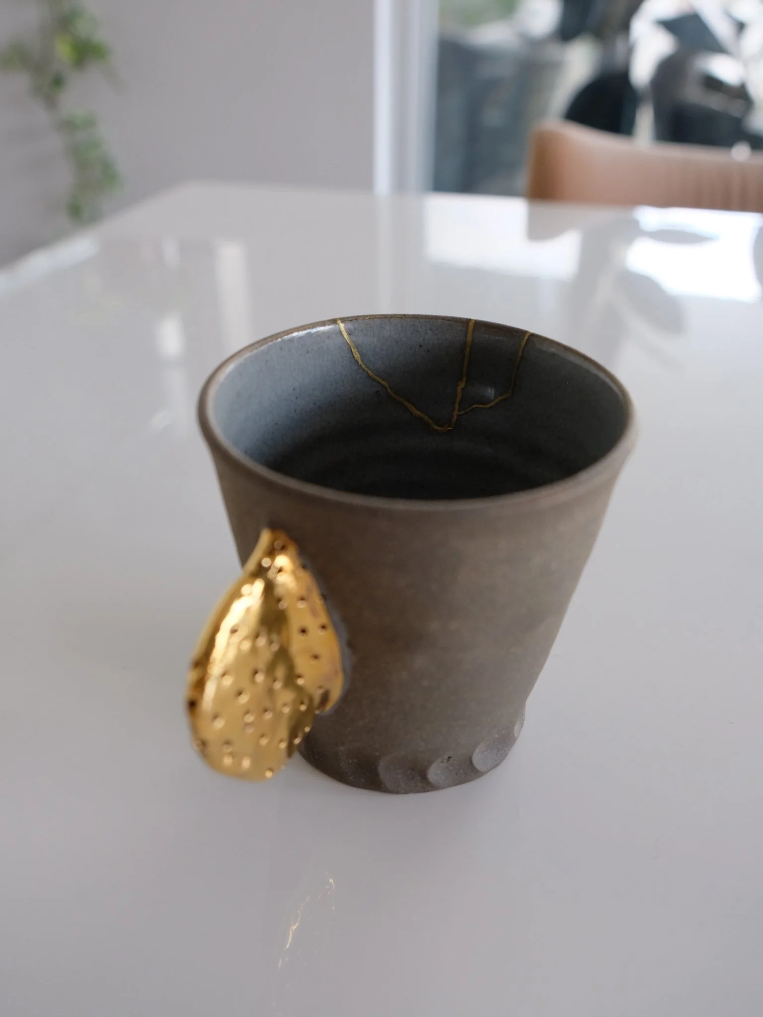 A gray ceramic cup with a gold leaf attached to the side, placed on a white surface, with a blurred background of a window and a chair. 23kt gold urushi food safe repair.