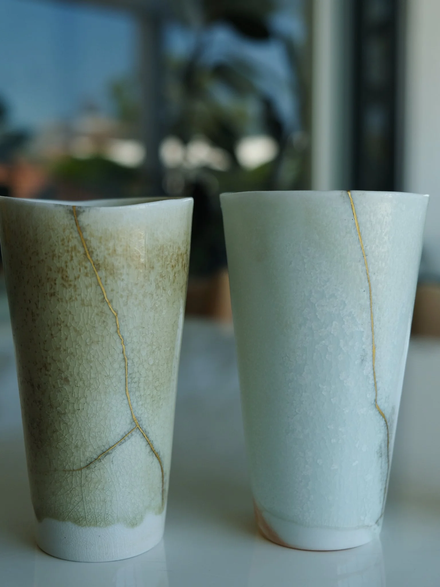 Two tall ceramic vases with crackle glaze and gold filigree, one in muted greenish and the other in light blue, placed on a white surface with a blurred background of indoor plants and windows.