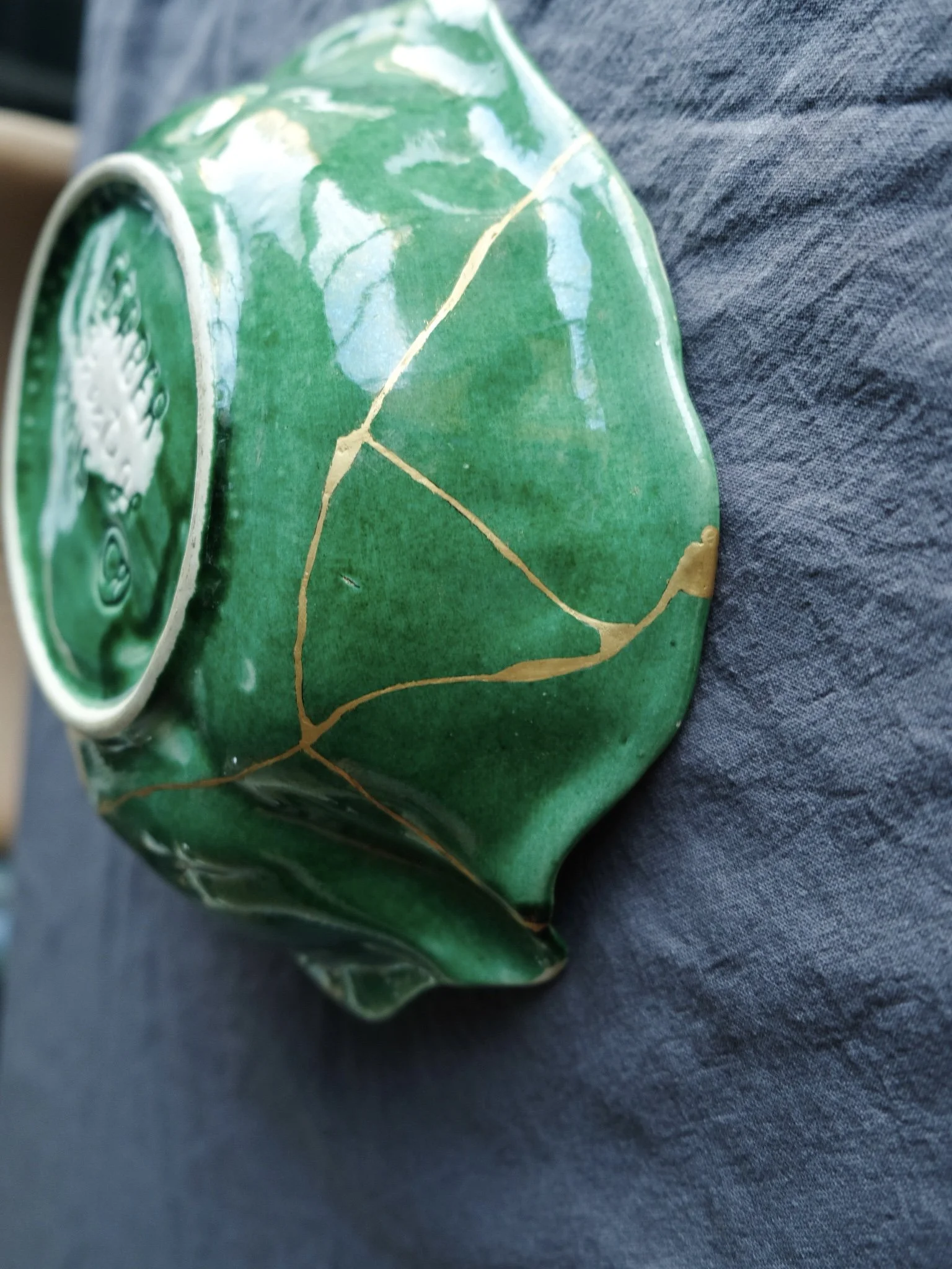A green ceramic bowl with gold-colored cracks and a decorative pattern, placed on dark fabric.