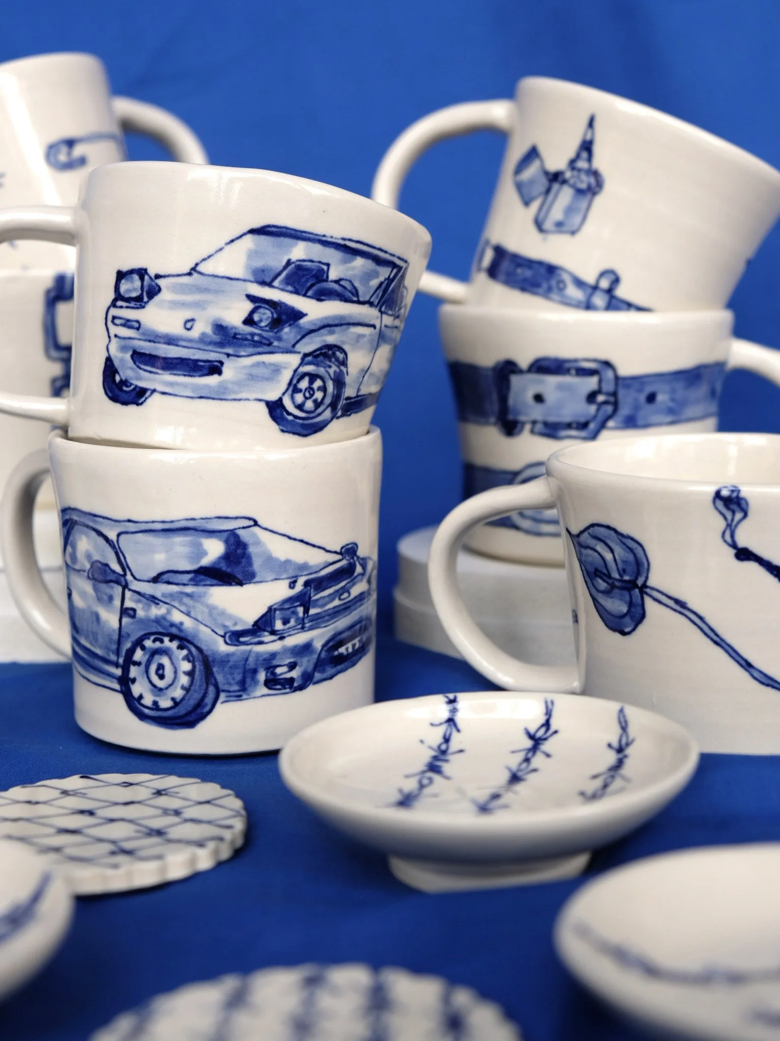 White ceramic mugs with blue illustrations of cars, belts, and a flower, arranged on a blue background.