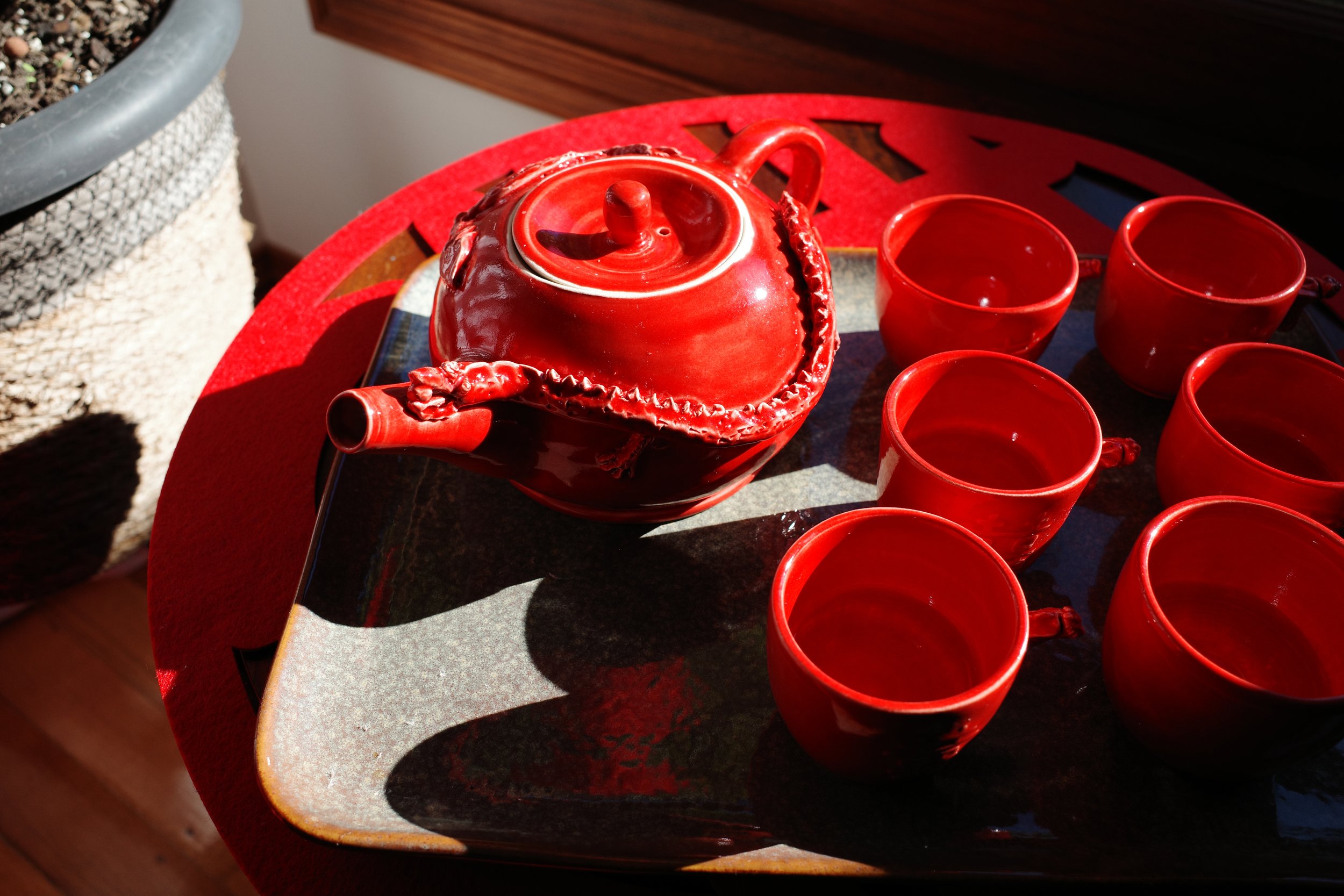 A red teapot with a decorative lid and spout, placed on a tray, alongside six red teacups, on a red table with sunlight casting shadows.