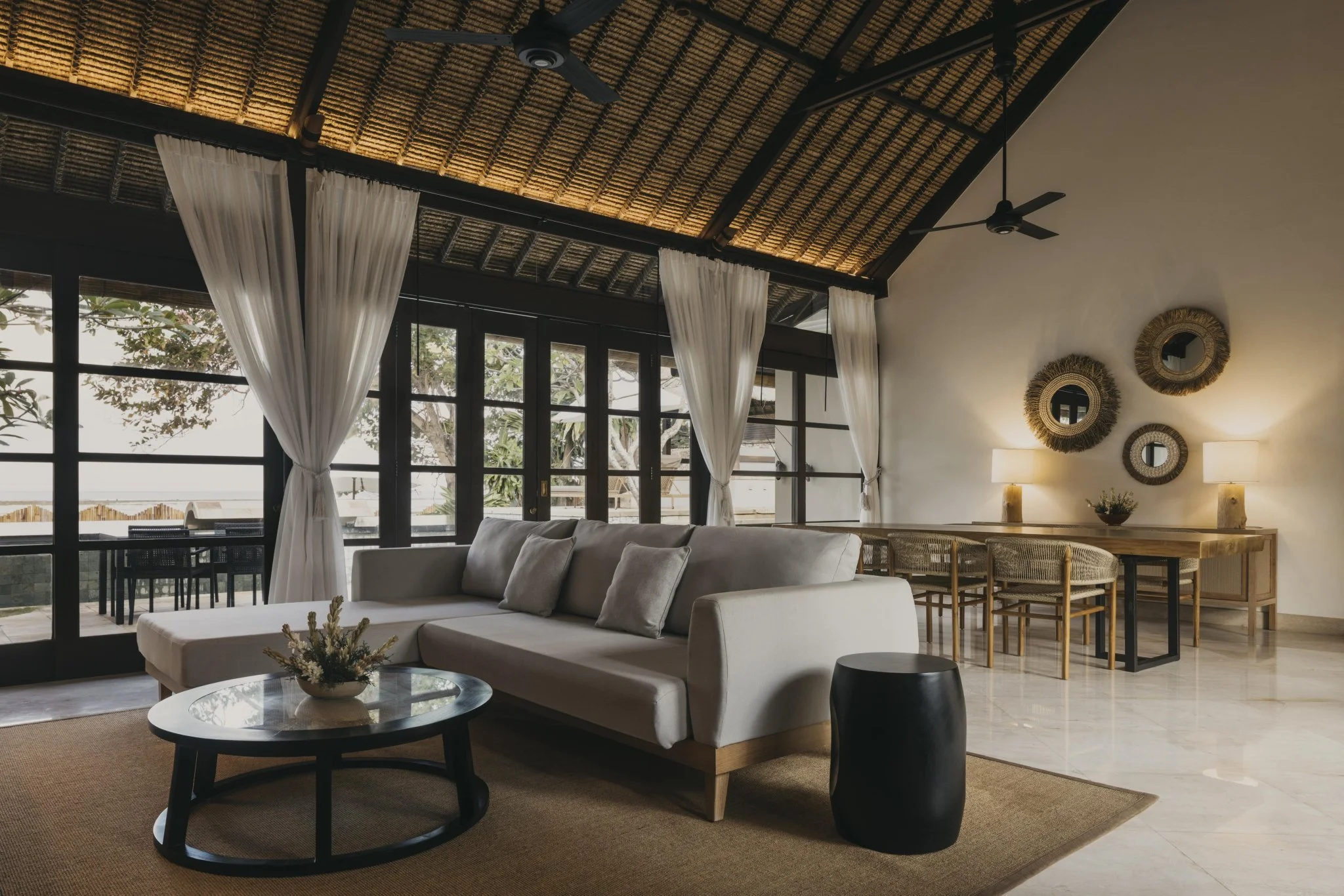 Modern tropical living room with large windows and soft white curtains overlooking the Bali beachfront. Custom-made sofa and round coffee table are paired with locally sourced Balinese wall décor and lamps.