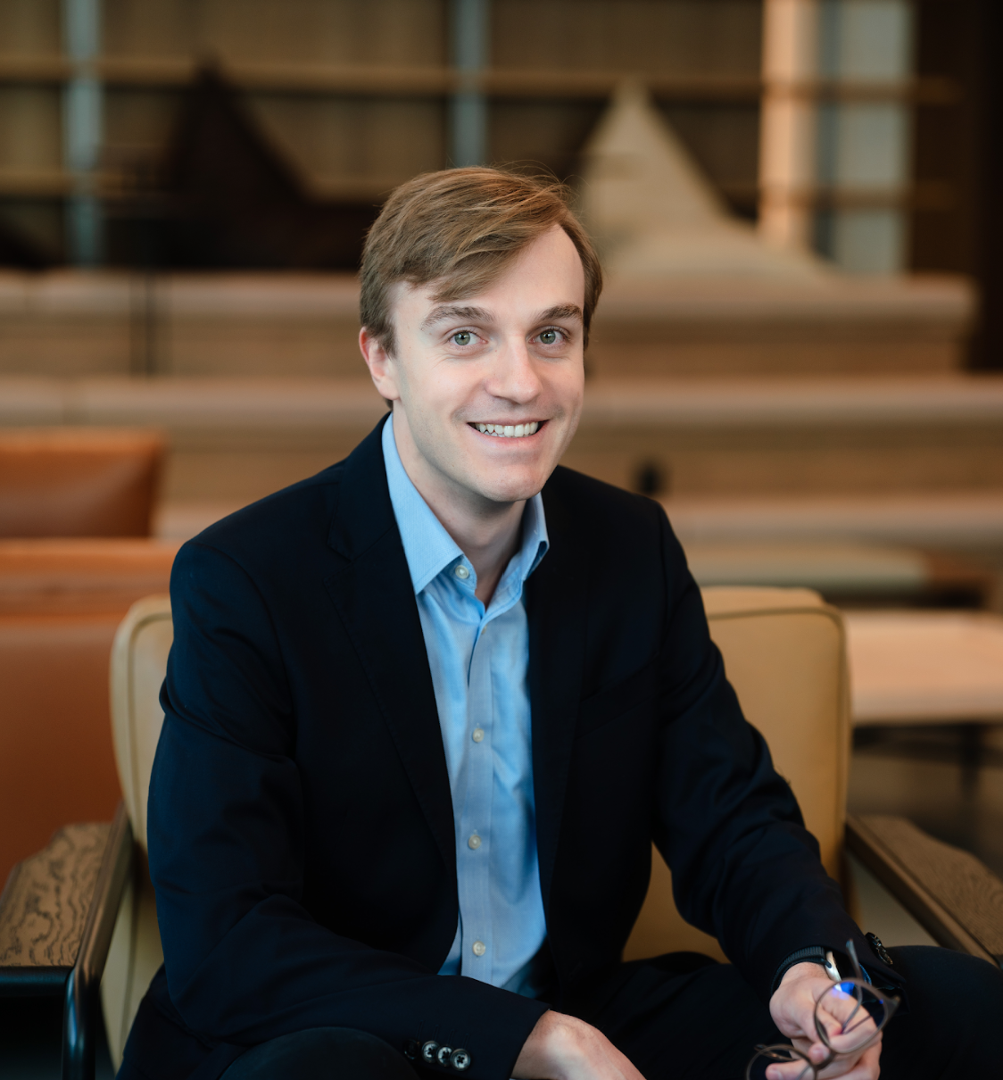 A young man in a dark suit and light blue shirt sitting on a chair in a modern indoor setting, smiling at the camera.