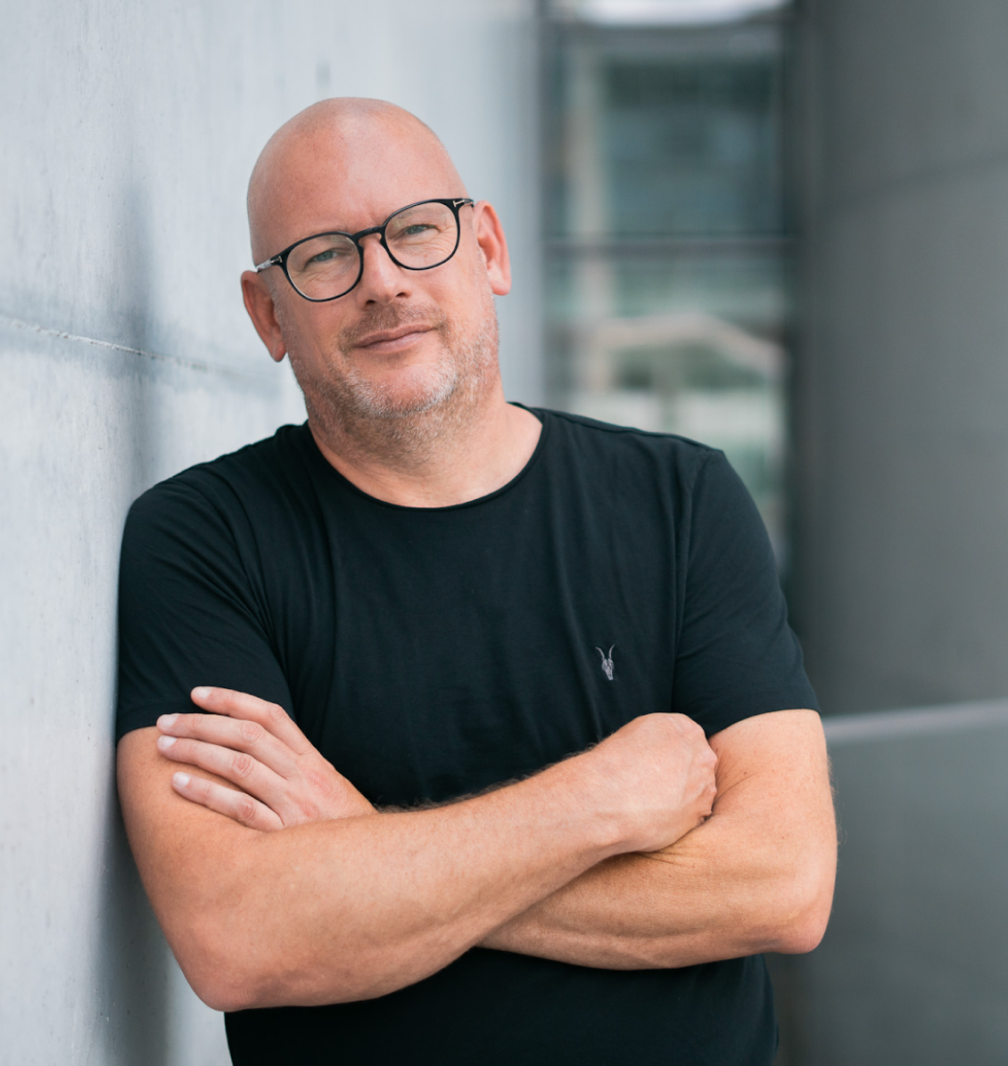 A middle-aged man with a bald head, glasses, and a short beard, wearing a black t-shirt, leaning against a gray concrete wall with arms crossed, smiling softly.