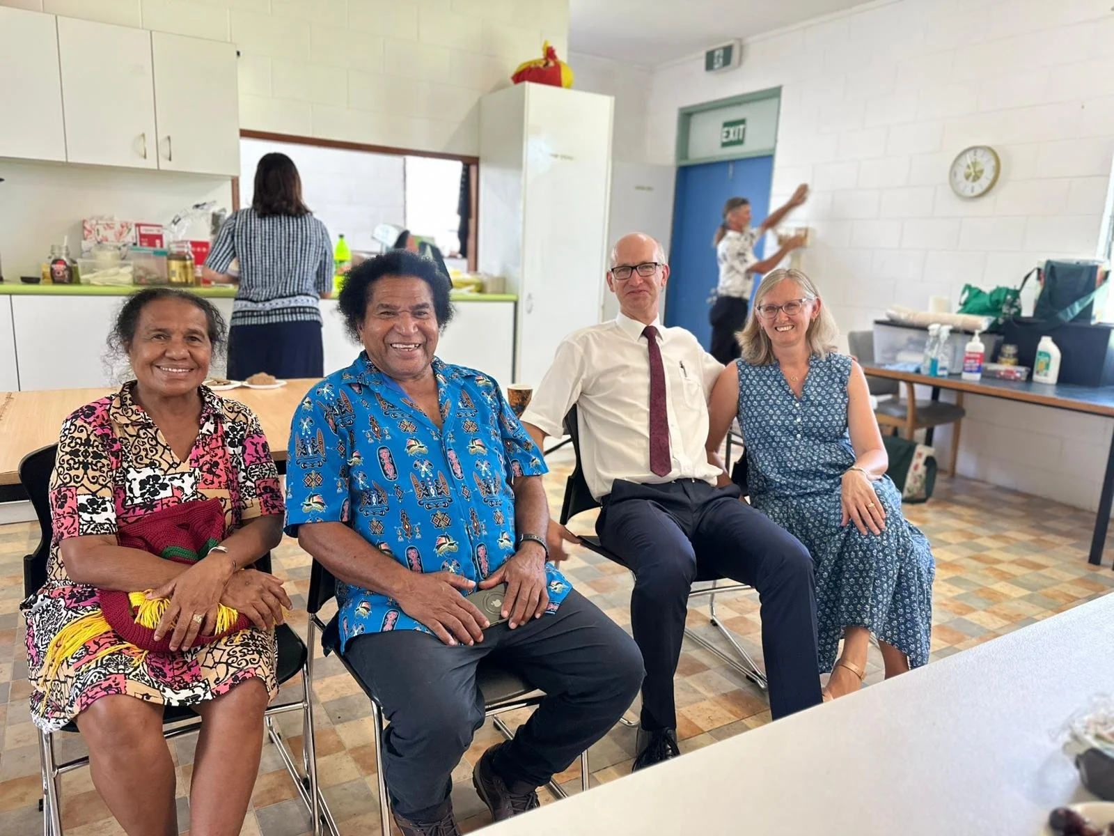 Four people at a church in Cairns, Australia, sitting and smiling at a table in a room with a kitchen area in the background, with two women and two men, all appearing happy and relaxed.