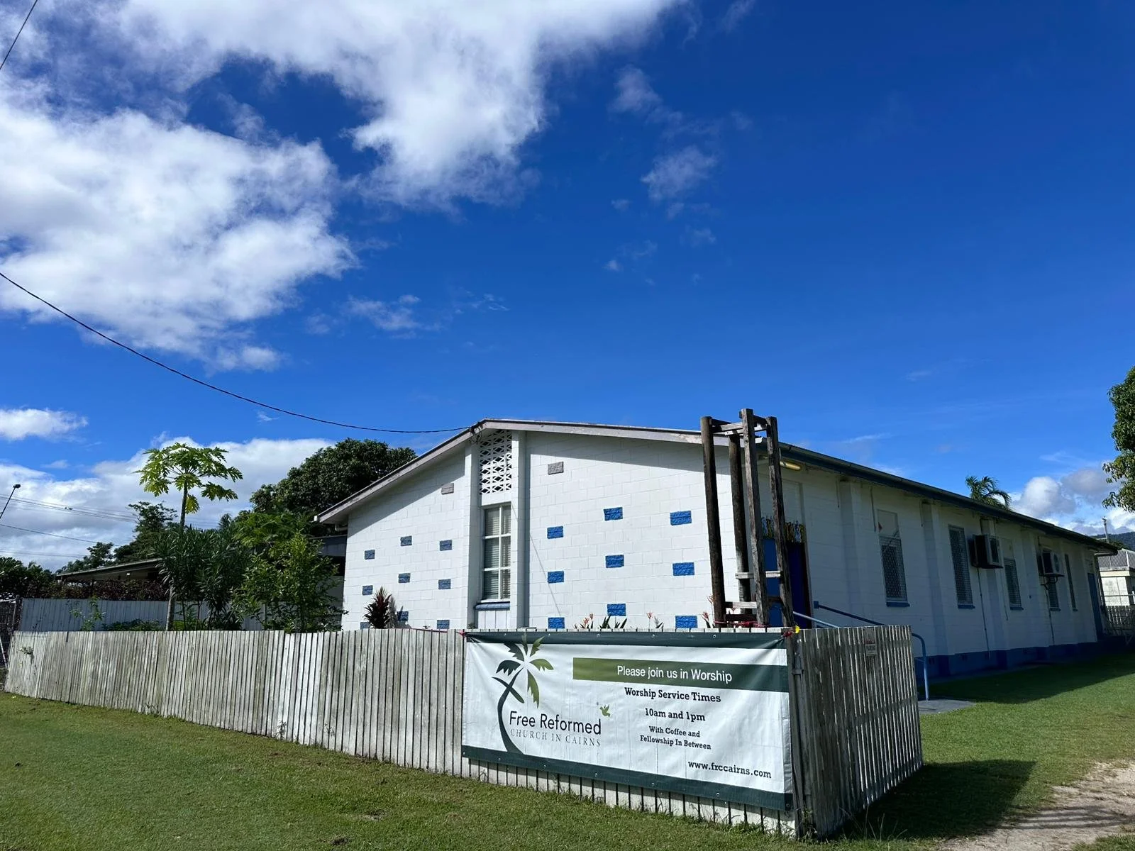 A white building with blue accents under a blue sky with clouds, surrounded by a wooden fence with a sign advertising worship service times at Free Reformed Church in Cairns, which is part of the Free Reformed Church of Australia