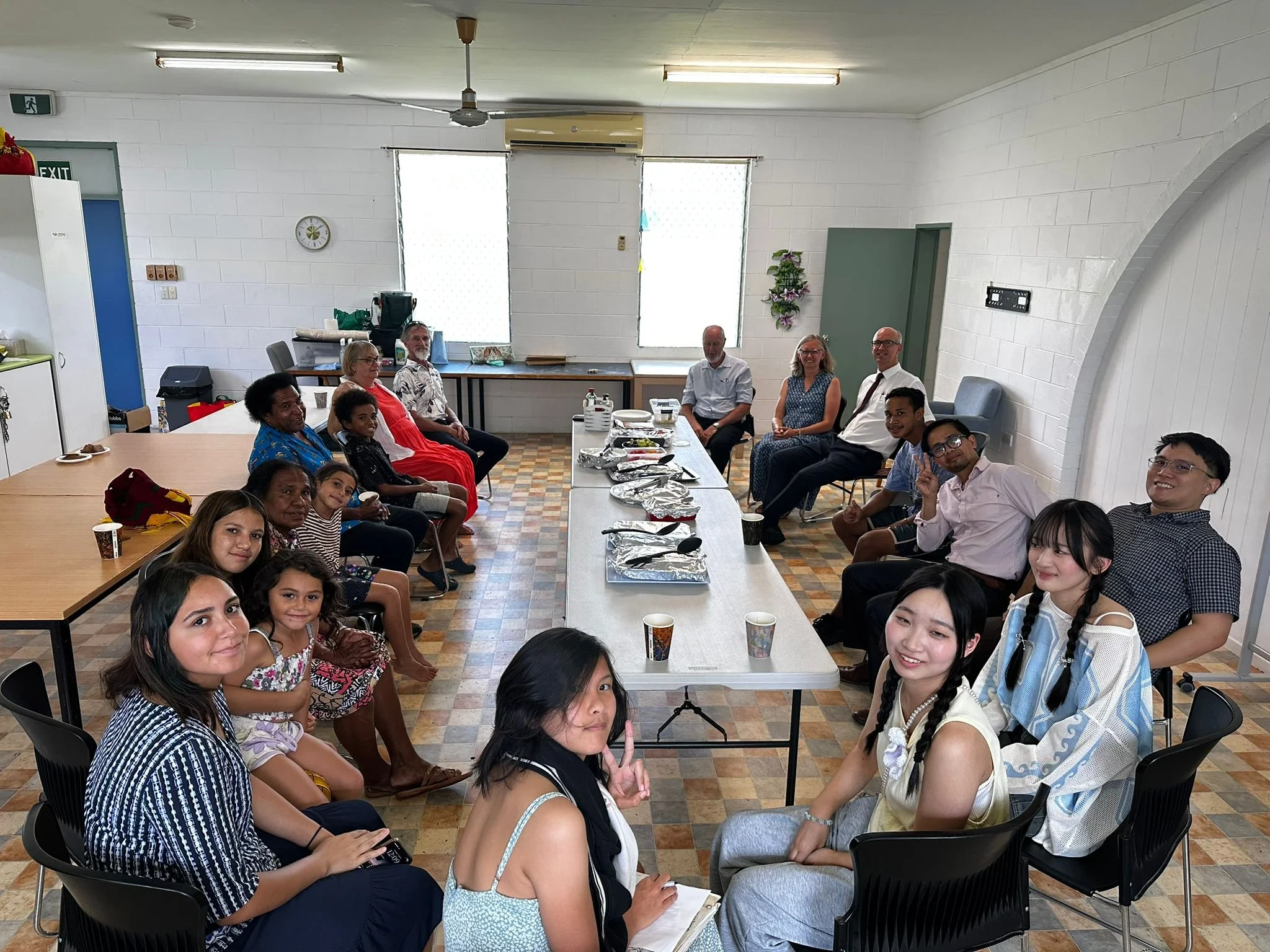 A diverse group of people, including children, teenagers, and adults, sitting around tables in a well-lit room at a church in Cairns, Australia, having a meal together. The tables are set with foil-covered dishes and cups