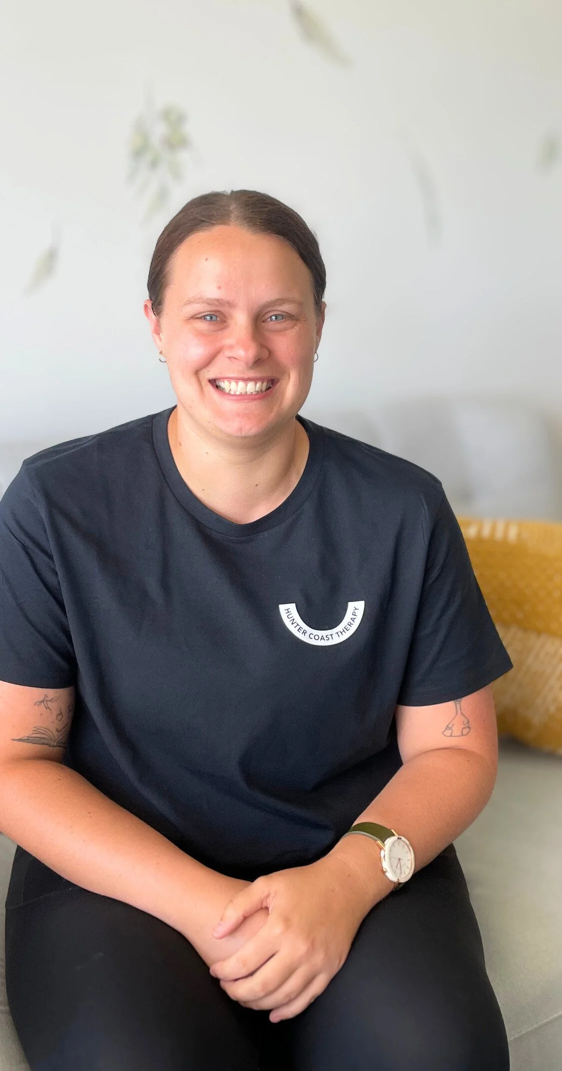 A smiling woman with short brown hair, wearing a black T-shirt with 'Hunter Coast Therapy' logo, sitting indoors against a white wall, with a yellow wicker basket in the background.