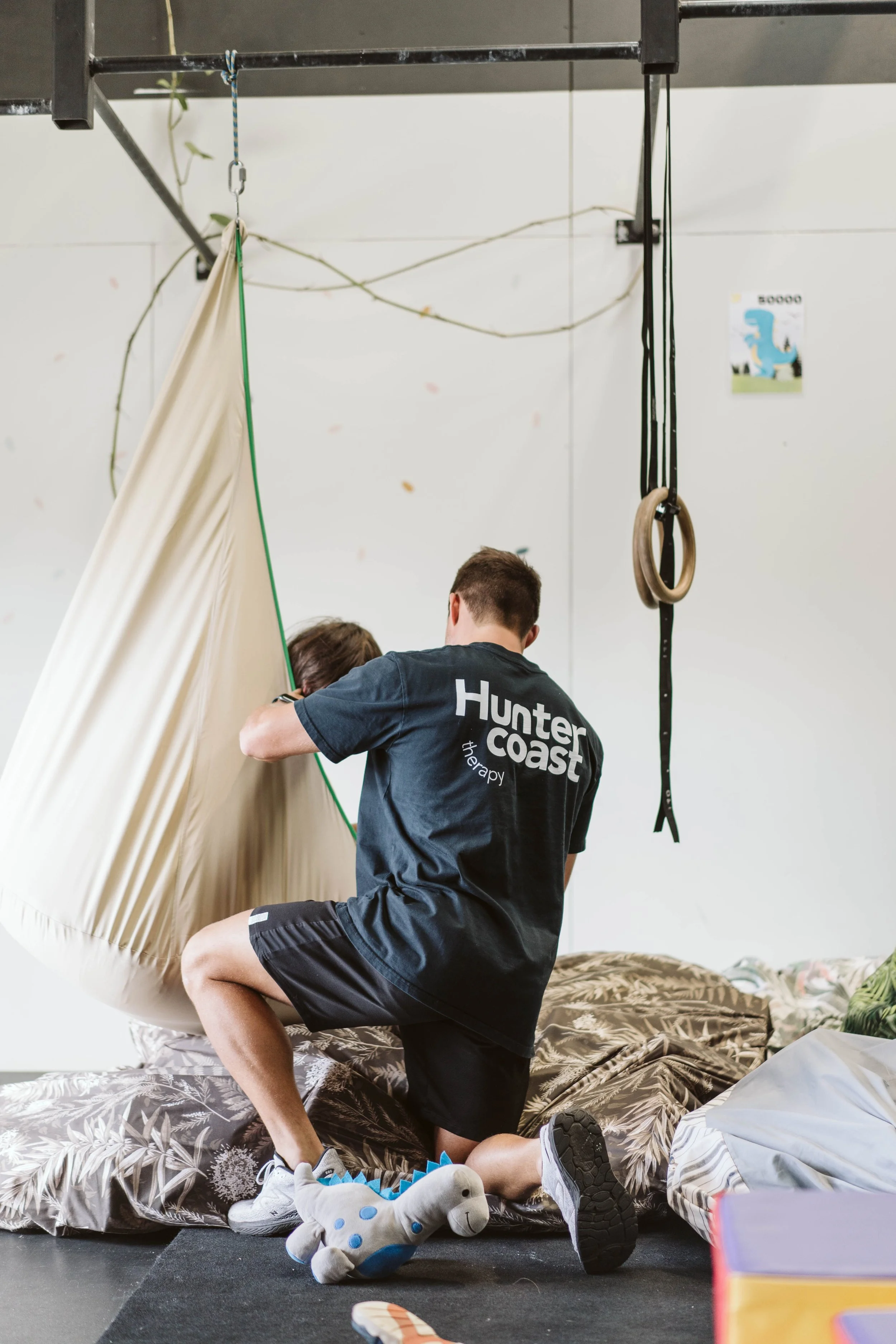 A therapy session in a gym with a therapist at Hunter Coast Therapy helping a young girl with a sensory swing. The room has minimal equipment, including gym rings and a poster of a blue dinosaur on the wall. A plush dinosaur toy is on the floor.