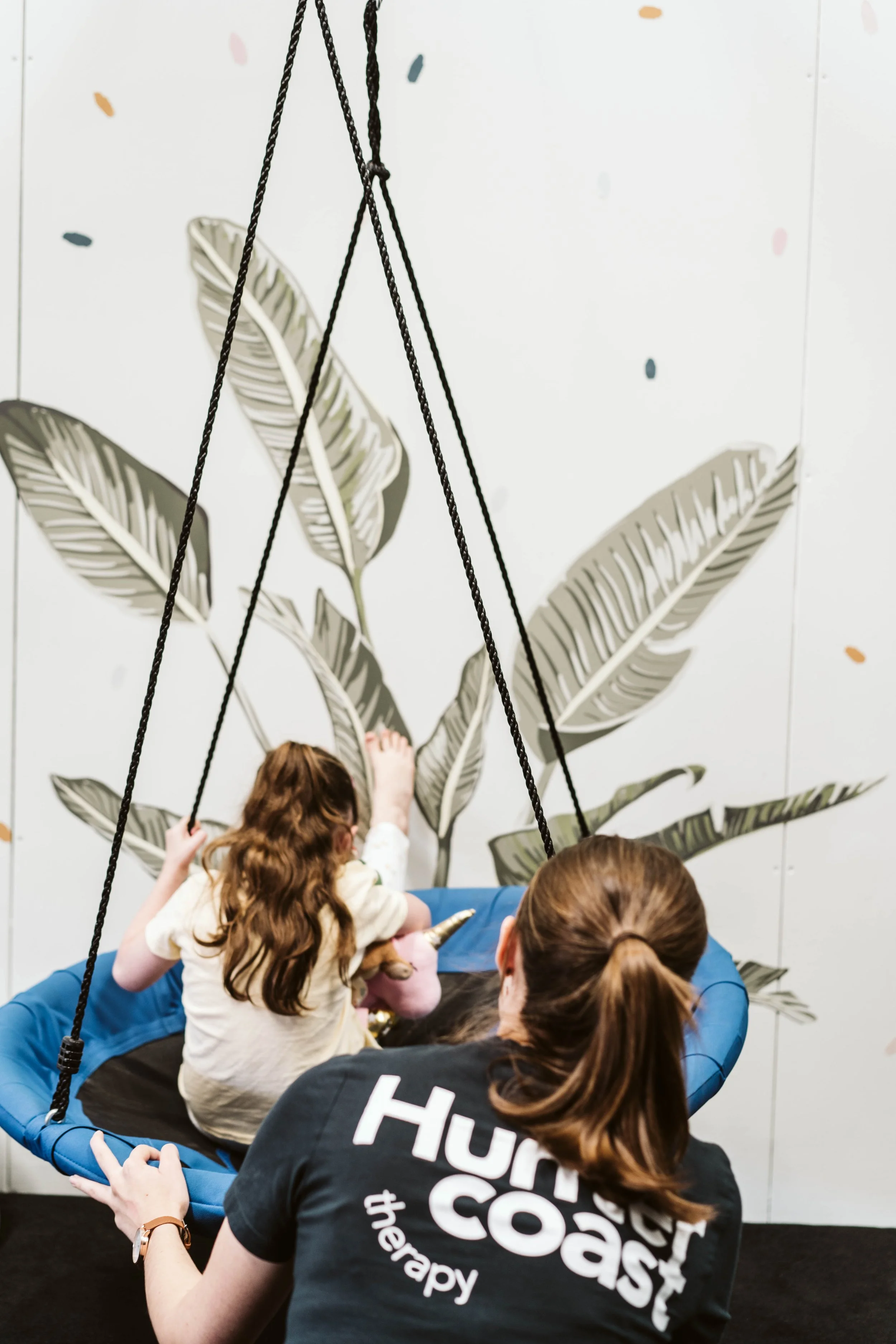 A young girl on a round swing with a blue cushion, sitting on a black surface, with an adult woman observing. The background features a mural of large, stylized leaves on a white wall. Newcastle Australia
