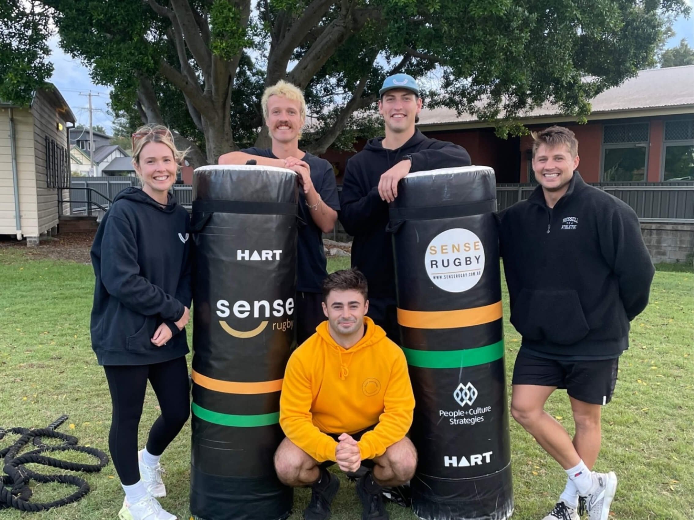 four men and a women standing around two rugby tackling bags in a park smiling
