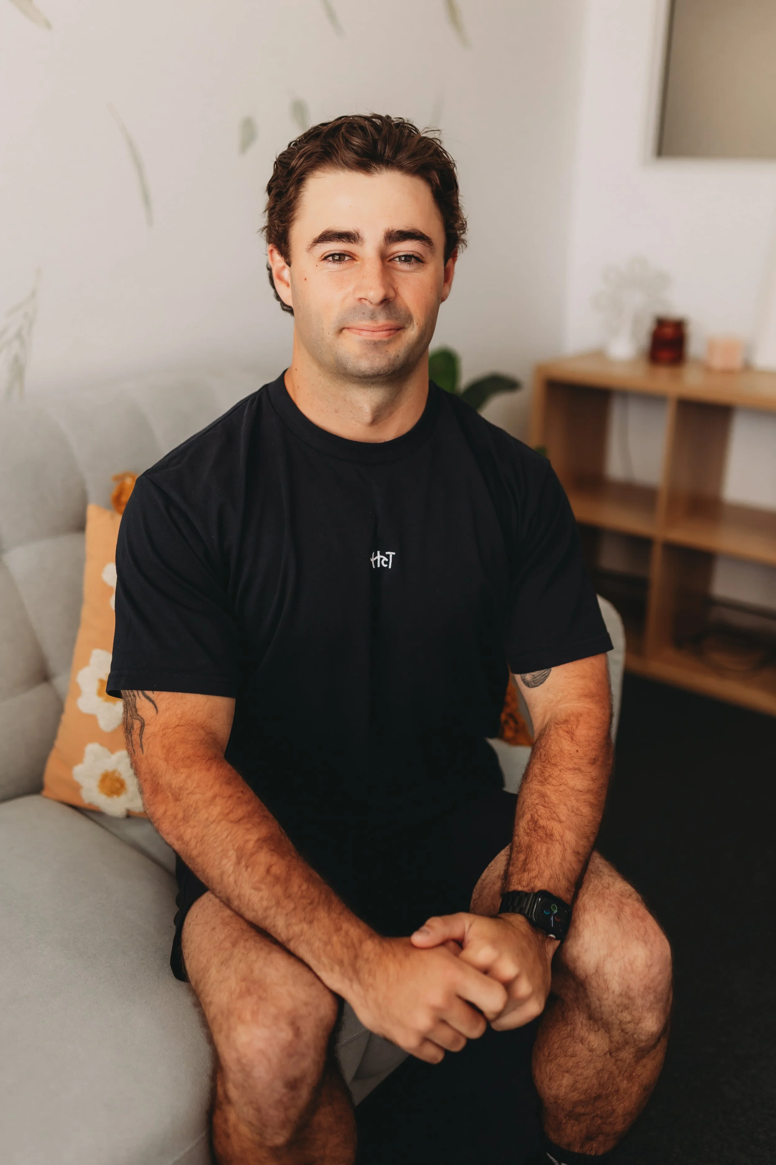 A young man with dark hair, wearing a black T-shirt and black shorts, sitting on a gray sofa with a floral pillow, in a cozy, modern living room. He is looking at the camera with a slight smile.