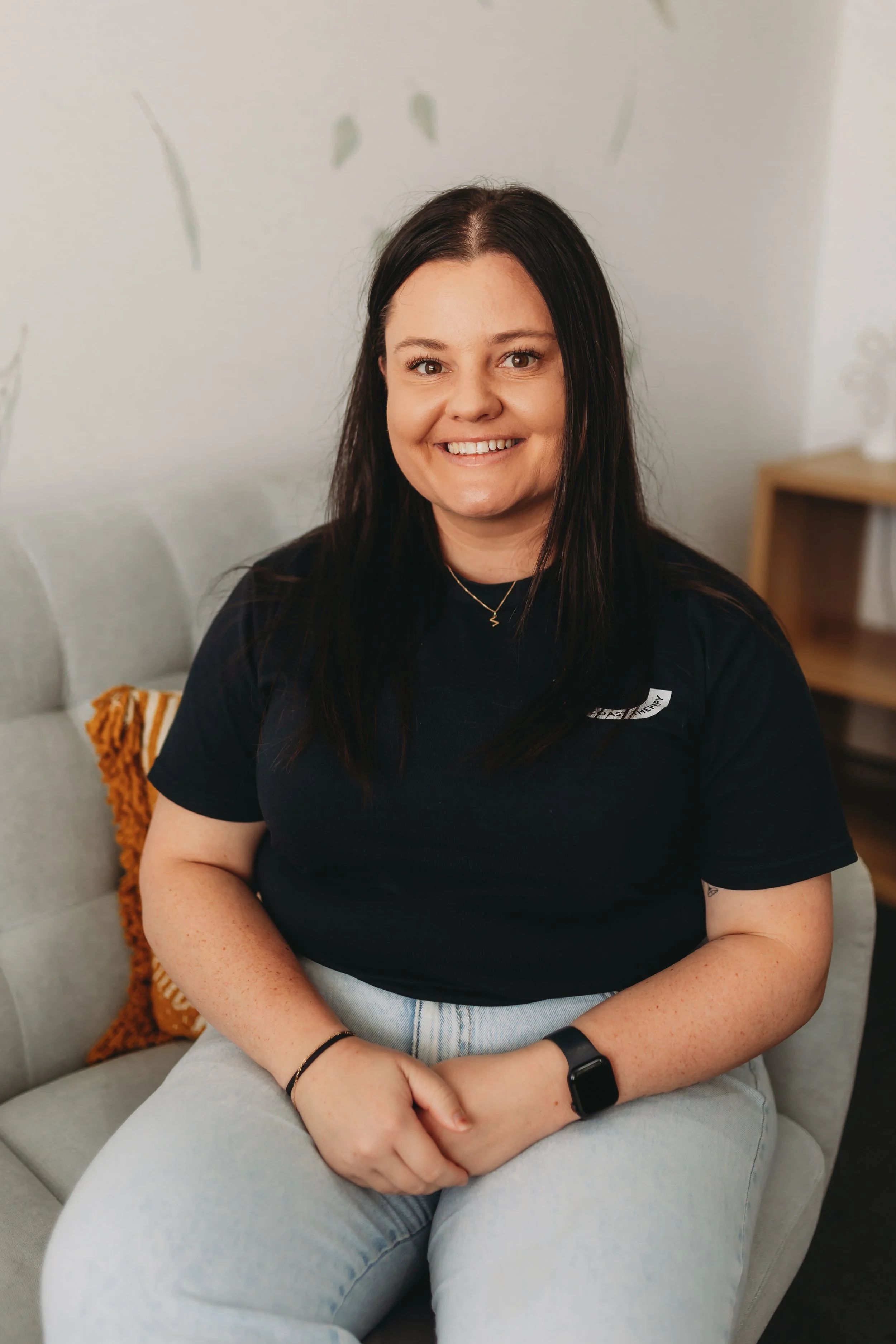 A woman with dark brown hair sitting on a light gray couch, smiling at the camera, wearing a black t-shirt, light jeans, a watch, and a necklace, with a decorative pillow behind her.