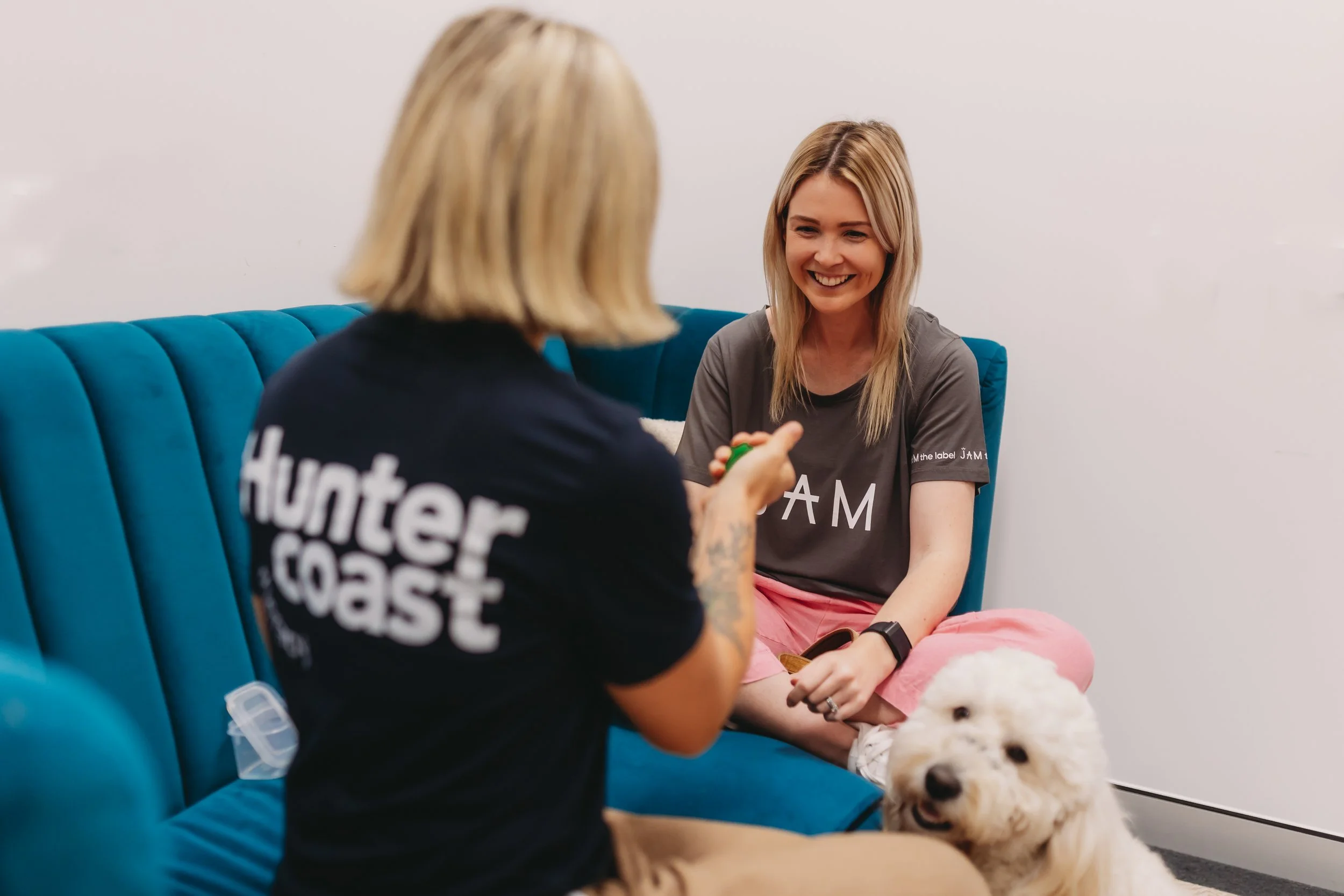 Two women with blonde hair sitting on a teal sofa, smiling and talking to each other, with a white dog sitting on the floor. Occupational Therapy Australia session.