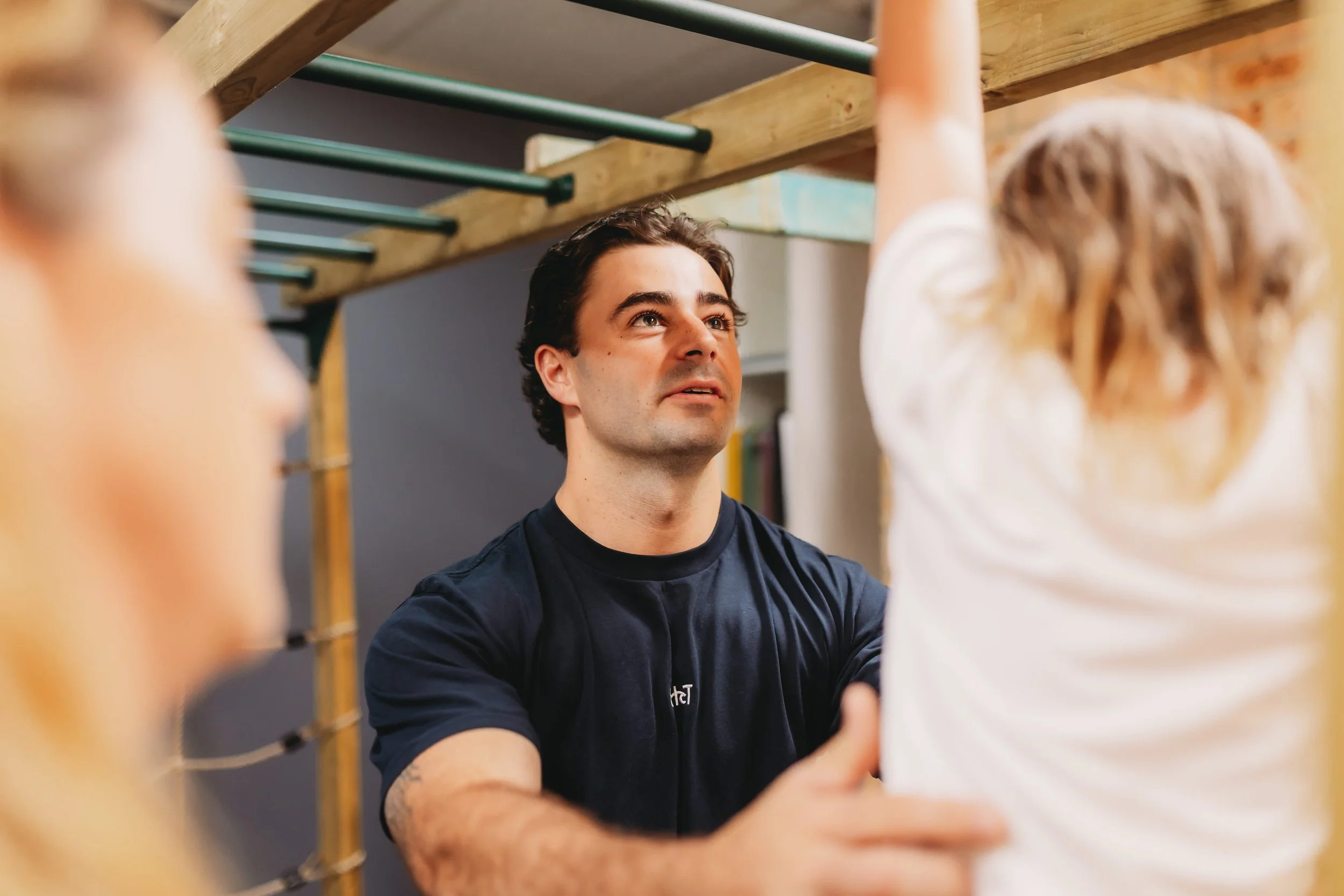 A man assisting a woman with physical therapy in a gym setting, with blonde woman in white shirt and a second woman in the foreground in Newcastle Australia