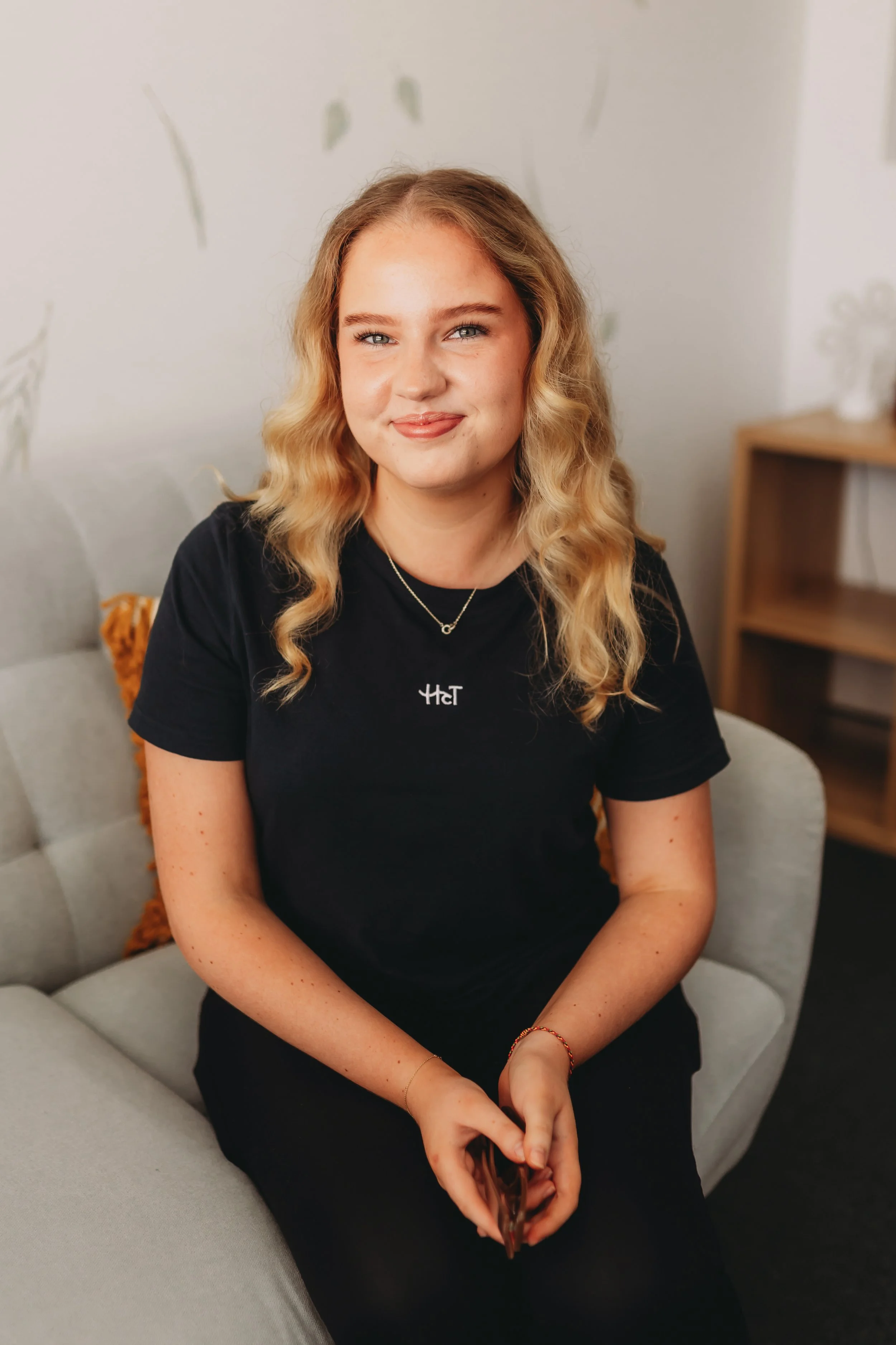 A young woman with blonde, curly hair sitting on a light gray couch in a living room, wearing a black T-shirt and holding a phone.
