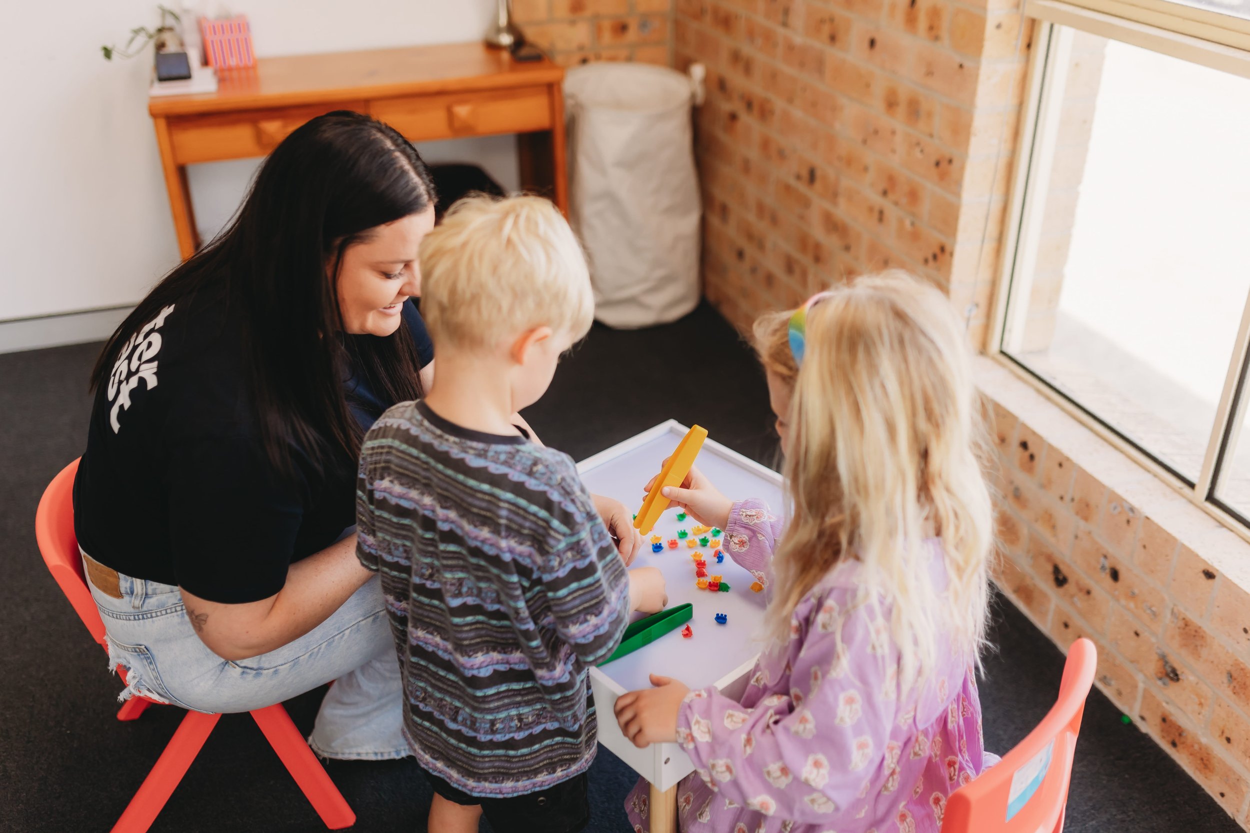 A woman and three children playing with small colorful toys in a cozy room with brick wall and large window. Occupational Therapy session Australia