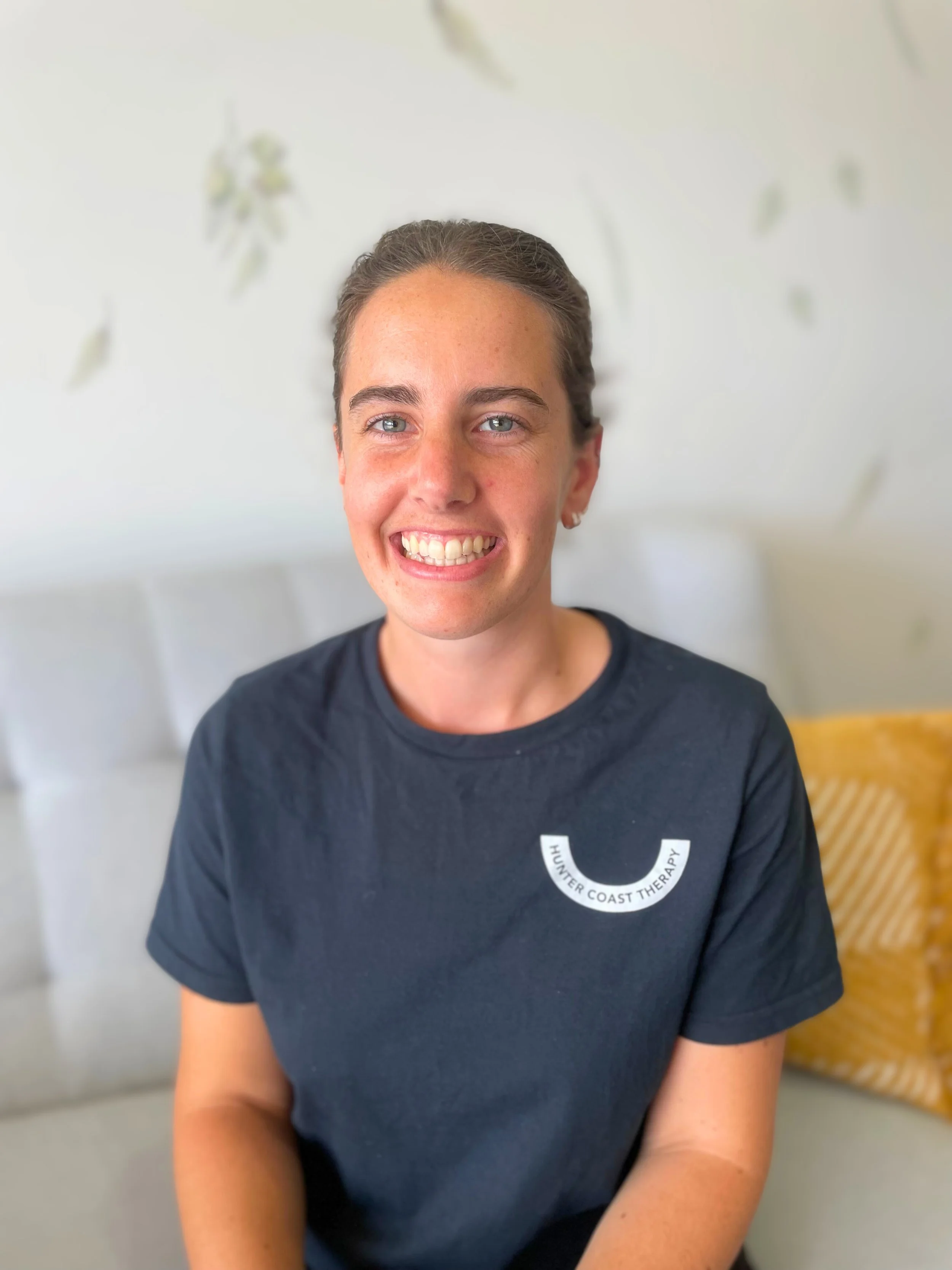 A young woman with brown hair tied back, smiling and showing teeth, wearing a navy blue shirt with a white logo that reads "Hunter Coast Therapy". She is seated on a light-colored couch with a yellow pillow behind her, in a room with a blurred white and green floral-themed wall.