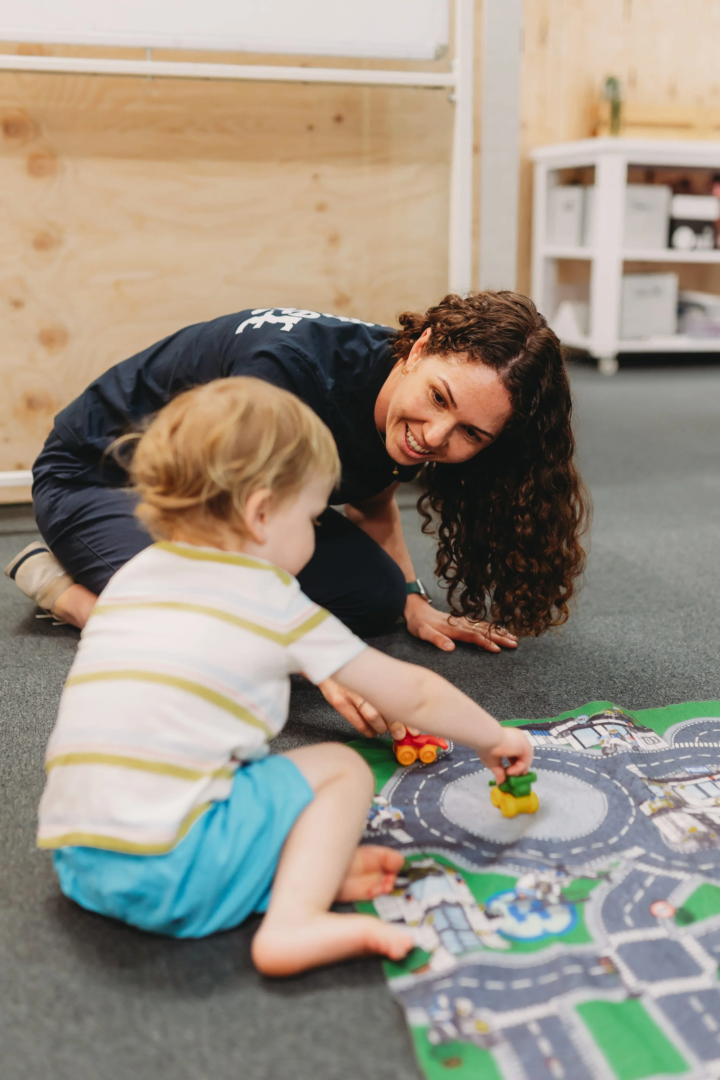 A woman and a young child playing with toy cars on a colorful play mat, indoors.