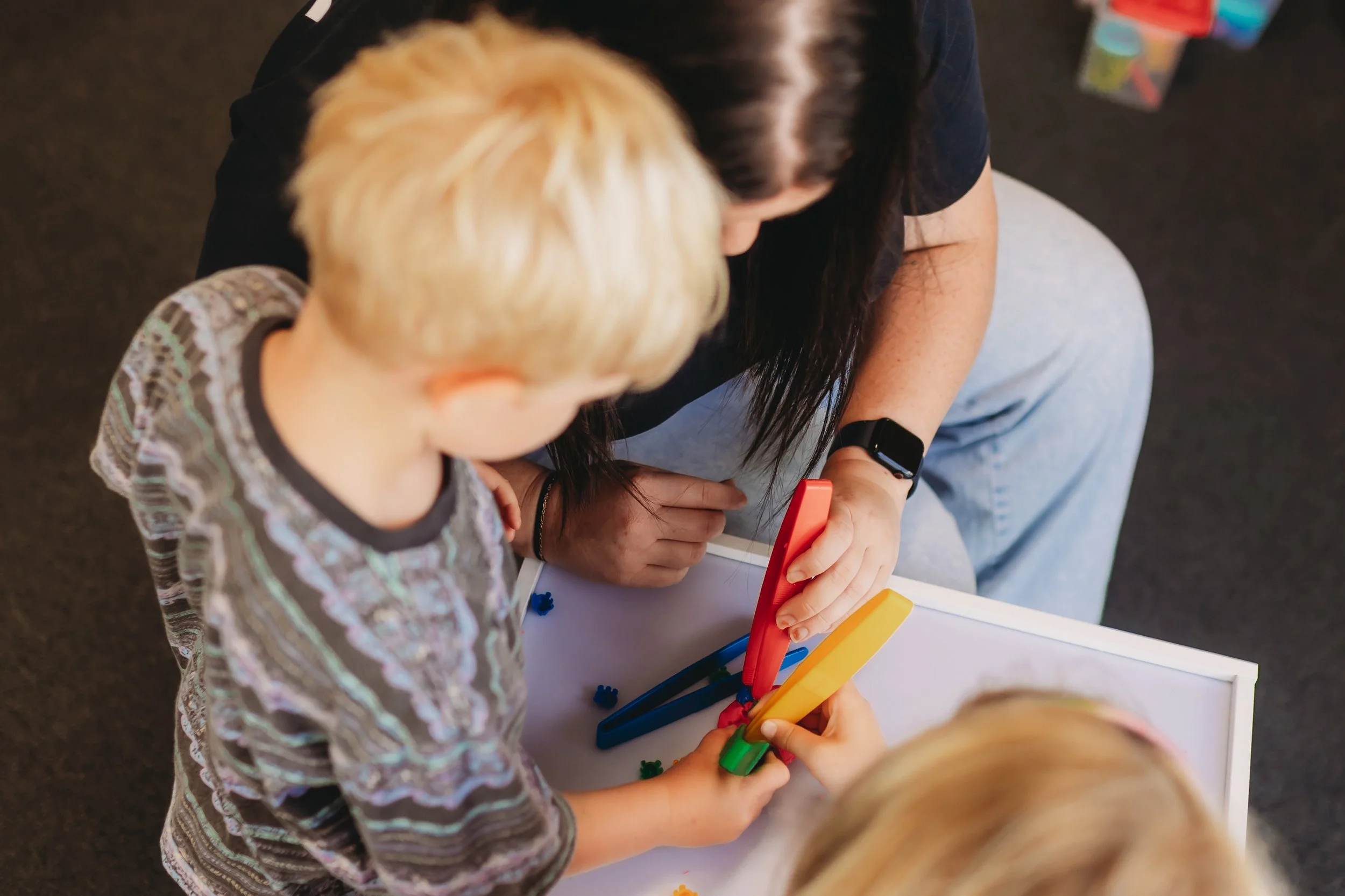 A woman and two young children with blonde hair playing with colorful magnetic sticks and shapes on a white surface in Newcastle Australia