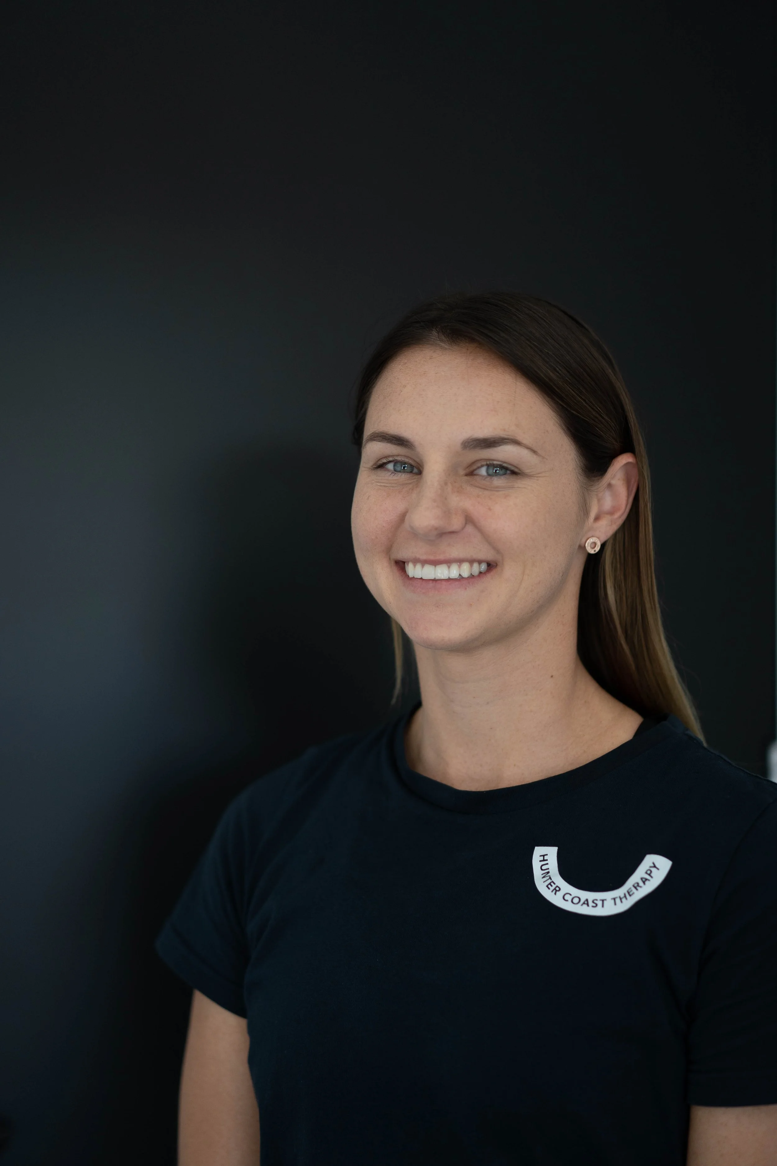 A young woman with long brown hair and blue eyes smiling, wearing a black shirt with a logo that reads 'HUNTER COAST THERAPY' in white text on a black background, standing against a dark backdrop.