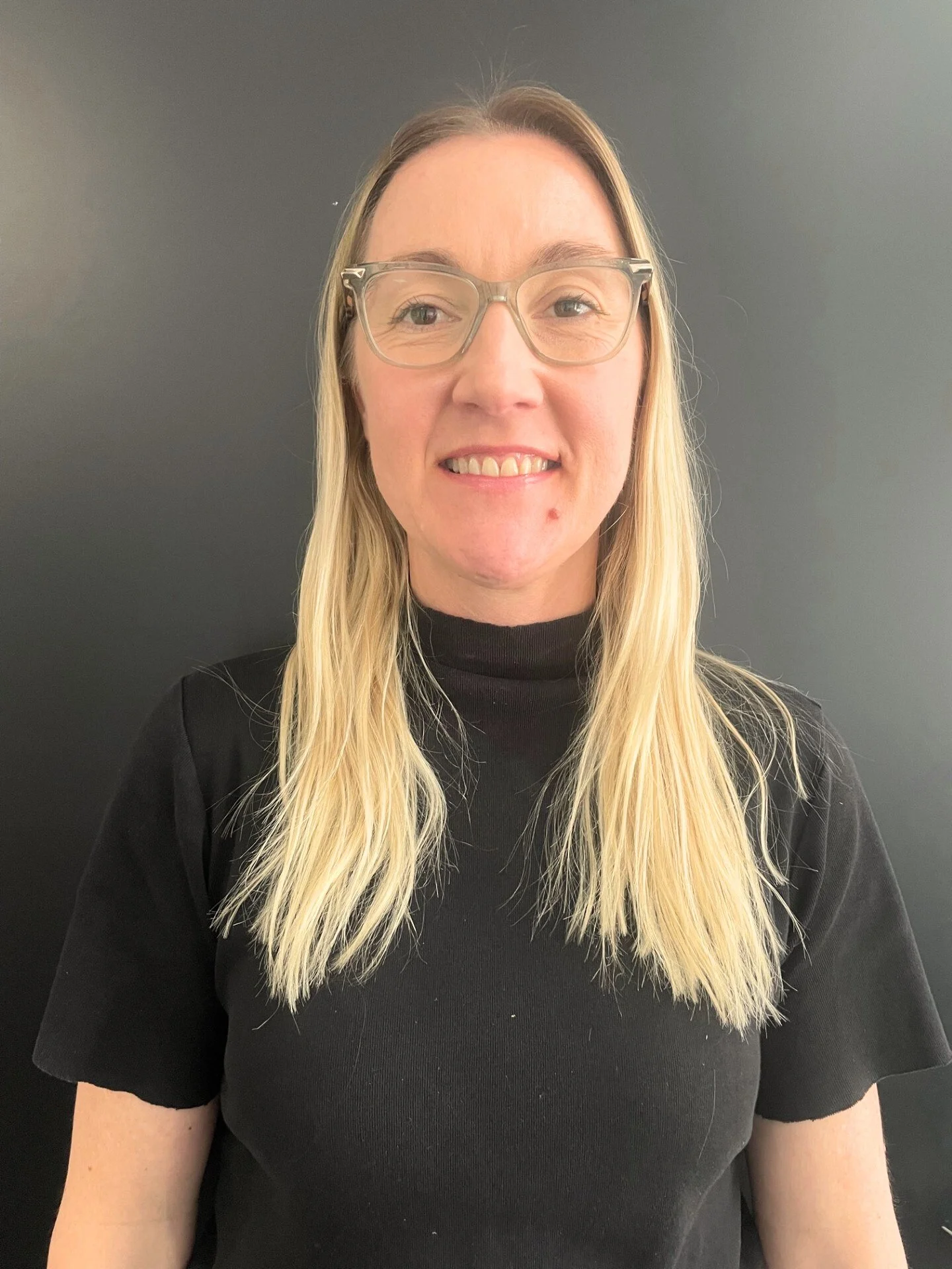 A woman with long blonde hair, glasses, and a black turtleneck shirt standing against a dark background.