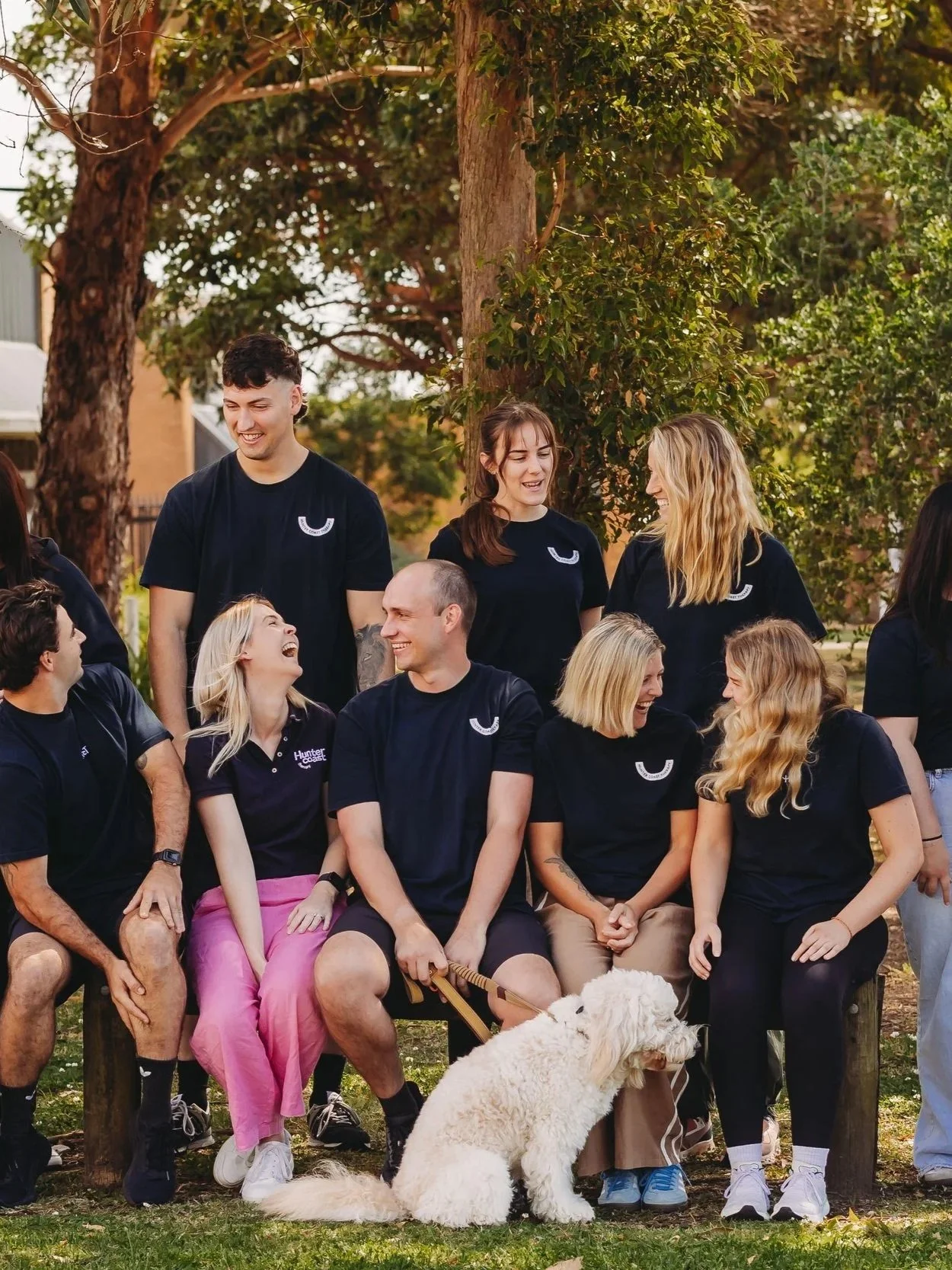 A group of people sitting outdoors on a bench and standing around, all wearing matching black shirts with a white logo, laughing and talking. There is a large tree in the background and a white dog sitting in front of the group.