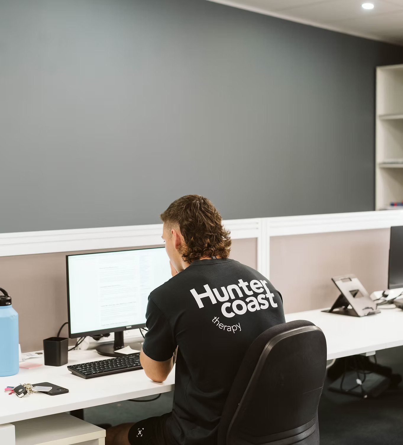 A person with long, curly brown hair sitting at a white desk in an office, working on a computer monitor. They are wearing a black T-shirt with white text that reads "Hunter coast therapy." There are keys, a phone, a water bottle, and other office supplies on the desk.