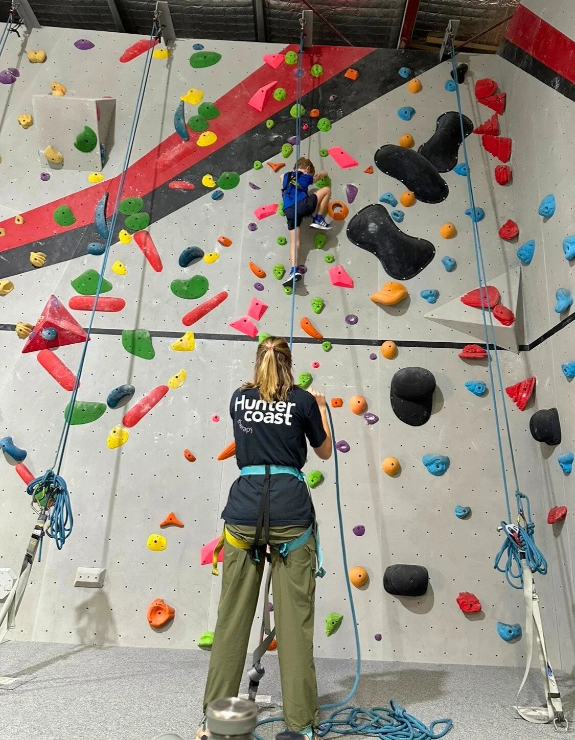An indoor climbing gym with colorful handholds and footholds. A young boy is climbing on the wall, and a woman called a belayer is standing at the bottom, managing the safety rope as part of therapy session in Newcastle Australia