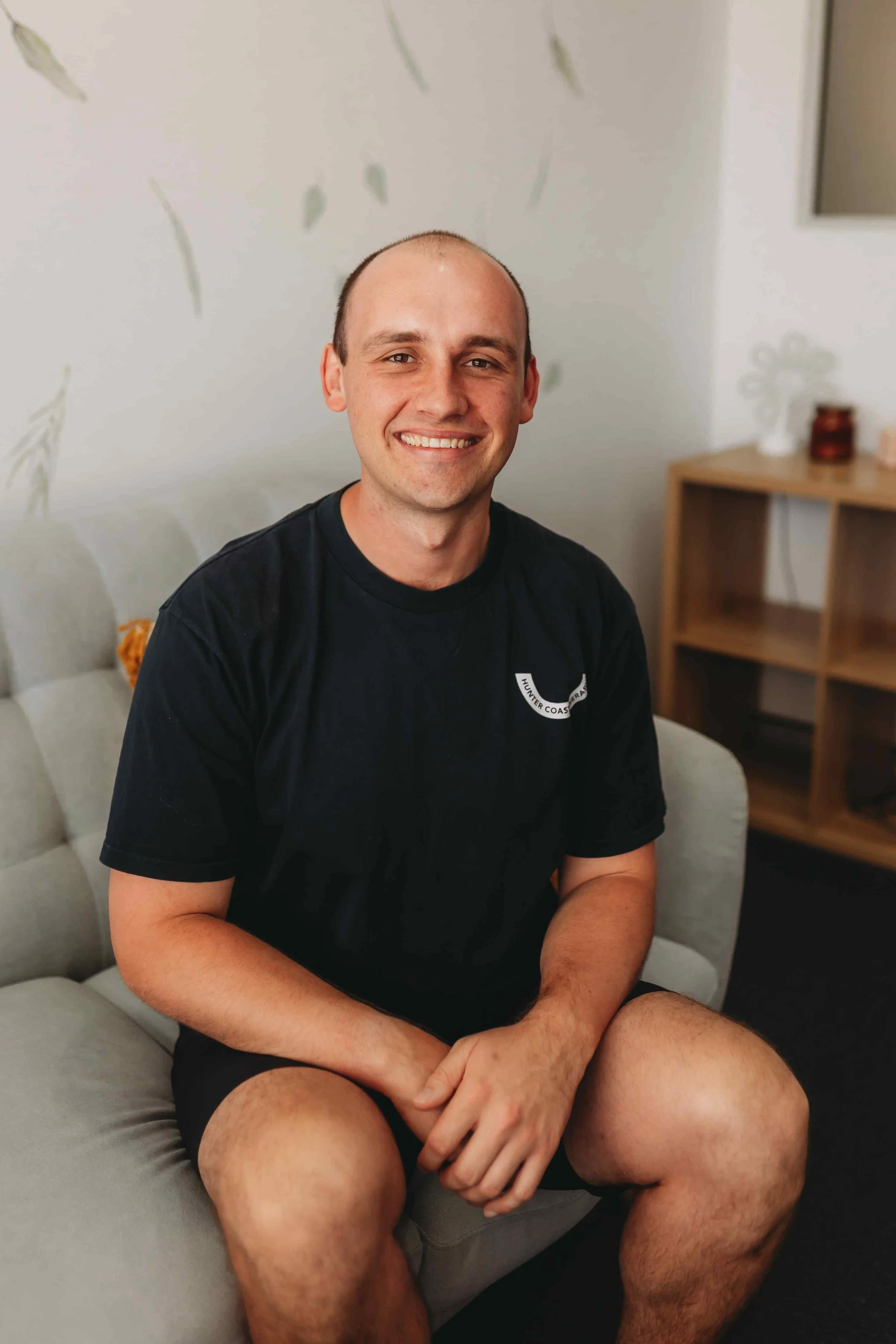A smiling man with a shaved head sitting on a sofa in a room with a light-colored wall and a wooden shelf in the background.