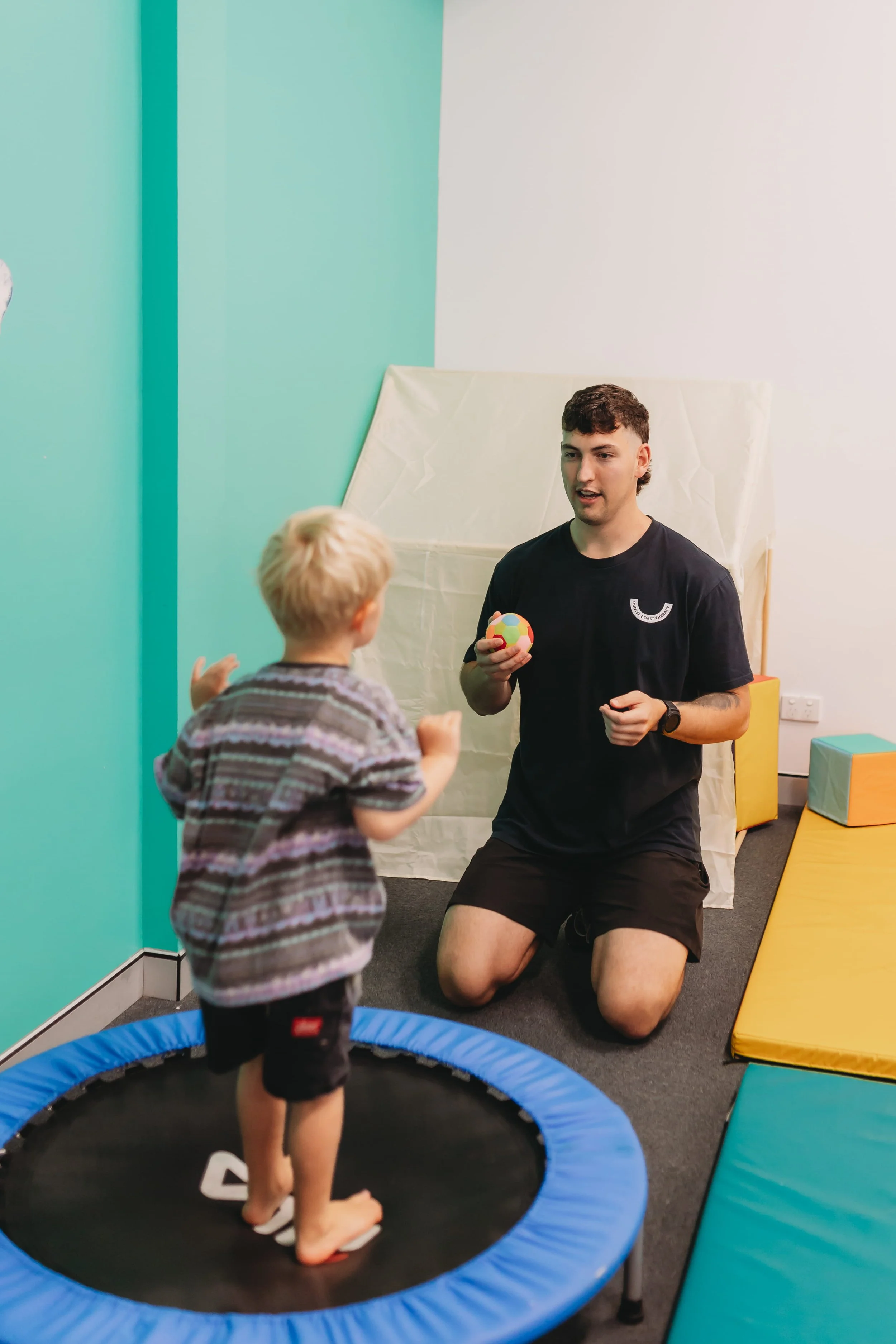 A young man kneeling on the floor talking to a child on a mini trampoline, while holding a colourful ball in a playroom with padded mats in Newcastle Australia