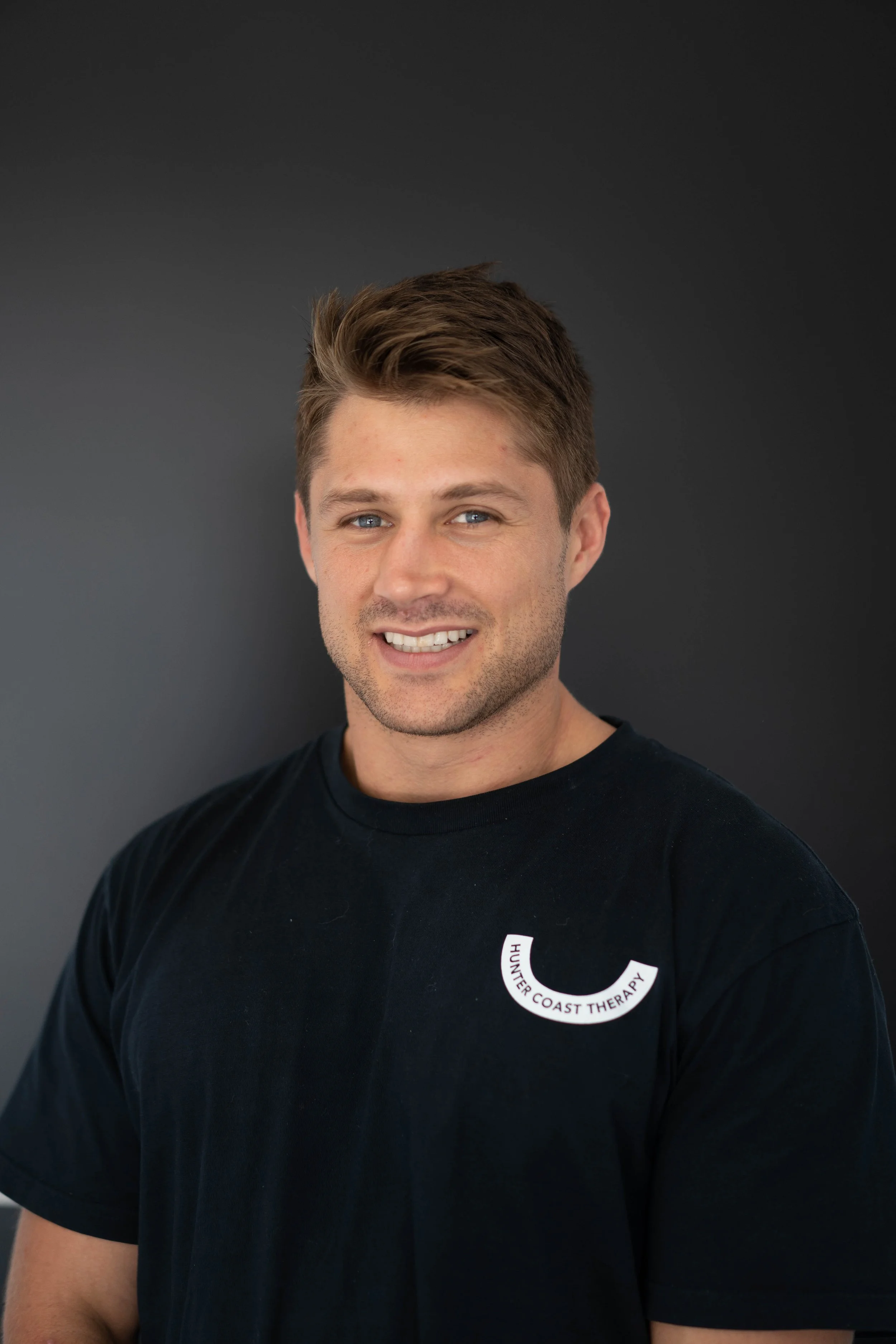 Headshot of a young man with brown hair, smiling, wearing a black T-shirt with a logo that reads 'HUNTER COAST THERAPY' on a black background.