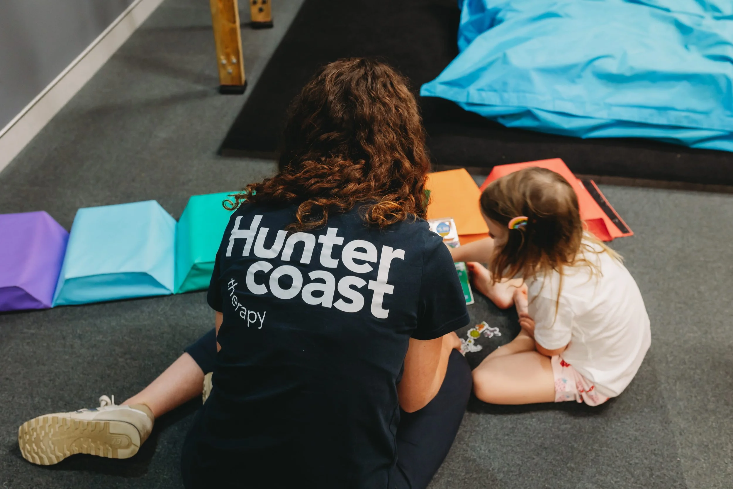 A woman with curly hair and a dark shirt that says 'hunter coast therapy' is sitting on the floor with a young girl, engaging in an activity with colorful books and toys. They are in a room with gray carpet, and there are colorful paper bags and large blue fabric in the background.