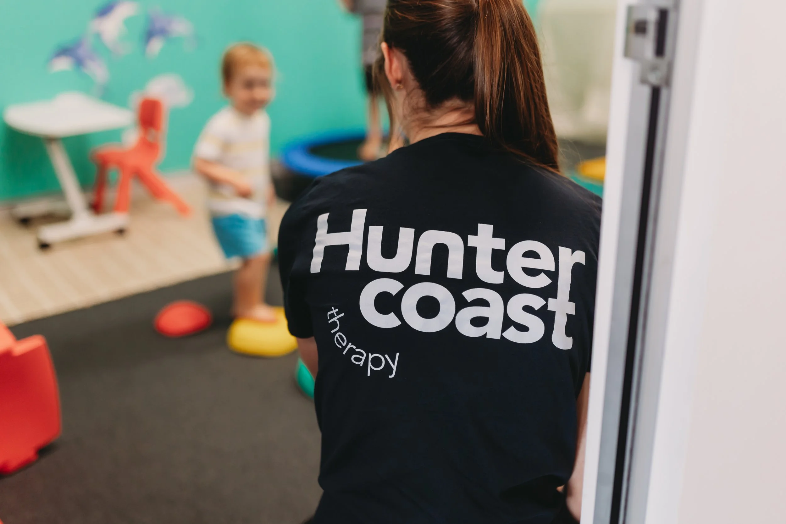 A therapy worker with 'Hunter Coast therapy' printed on her black T-shirt observes two children playing in an indoor therapy room with colorful equipment and a mural of dolphins on the wall.