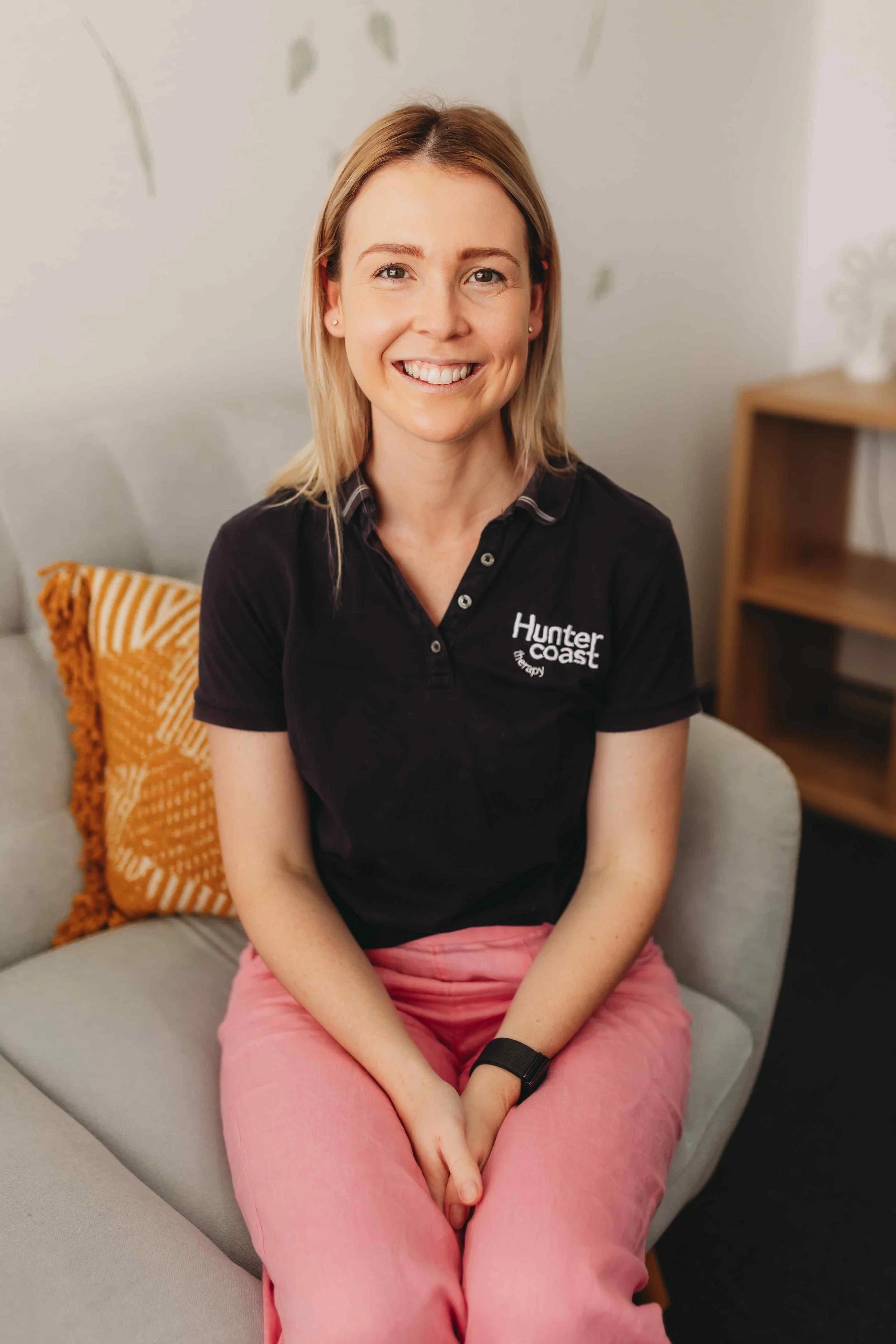 A woman with shoulder-length blonde hair smiling, sitting on a light-colored couch with an orange pillow behind her. She is wearing a black polo shirt with 'Hunter Coast' embroidered on it and pink pants.