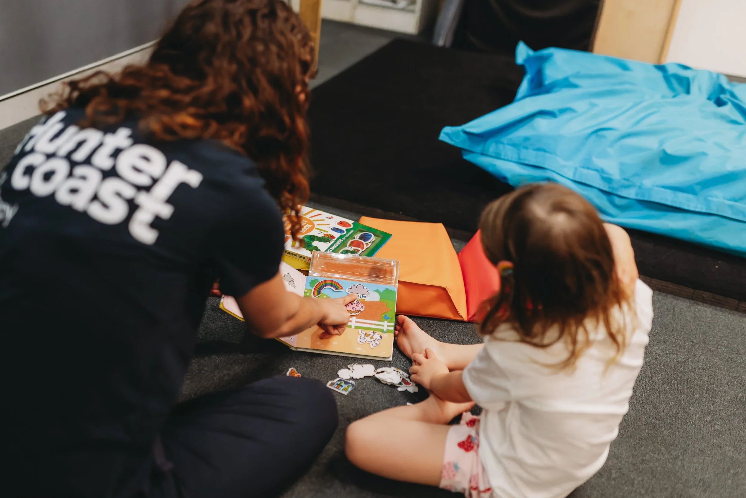 An adult and a young girl sitting on the floor engaging with children's books and stickers. The adult is wearing a shirt that says 'Hunter Coast' on the back. The girl has curly hair and is wearing a white t-shirt. OT Session Australia