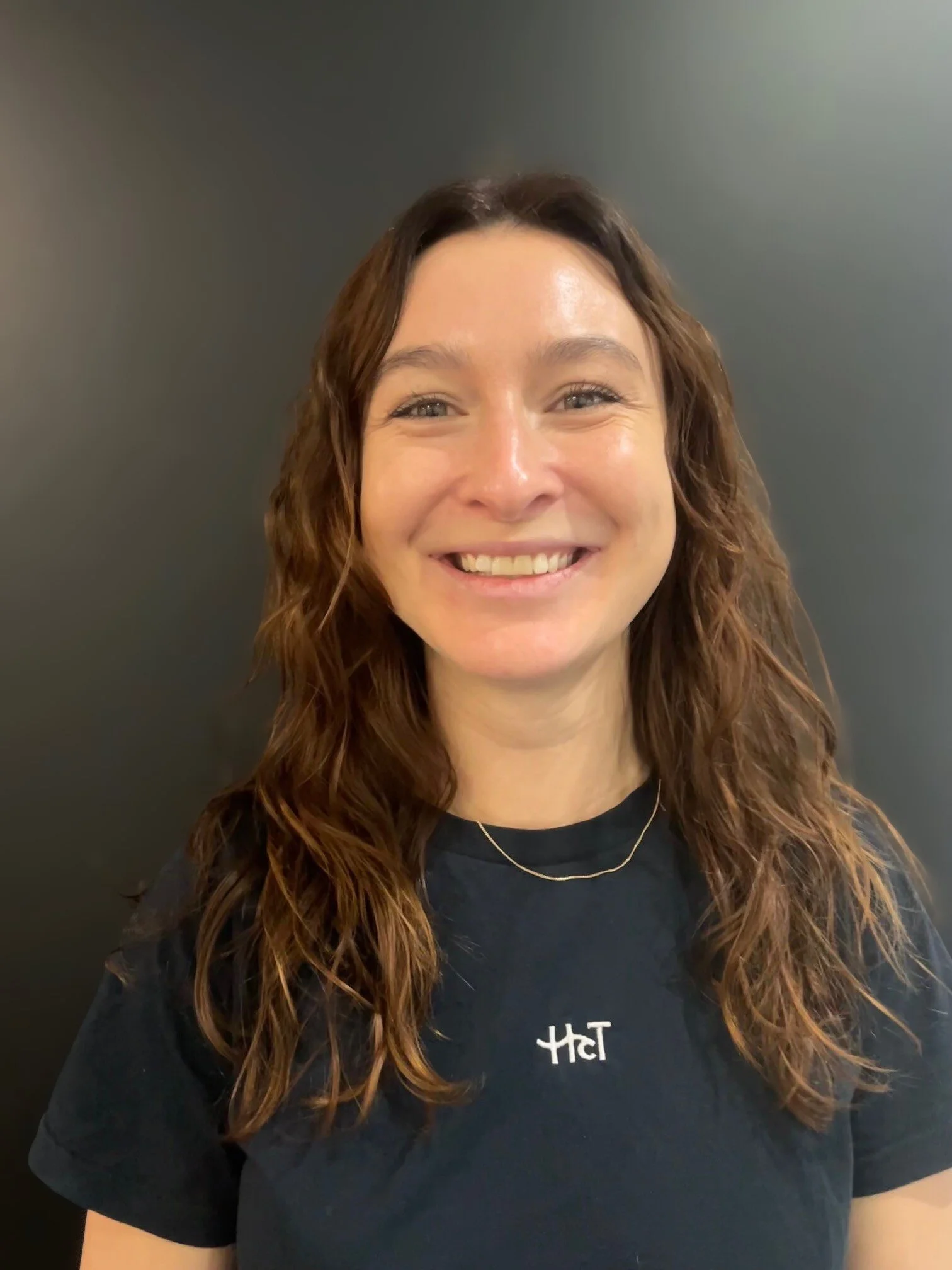 A woman with long wavy brown hair smiling, wearing a black T-shirt with white text and a gold necklace, standing against a plain dark background.