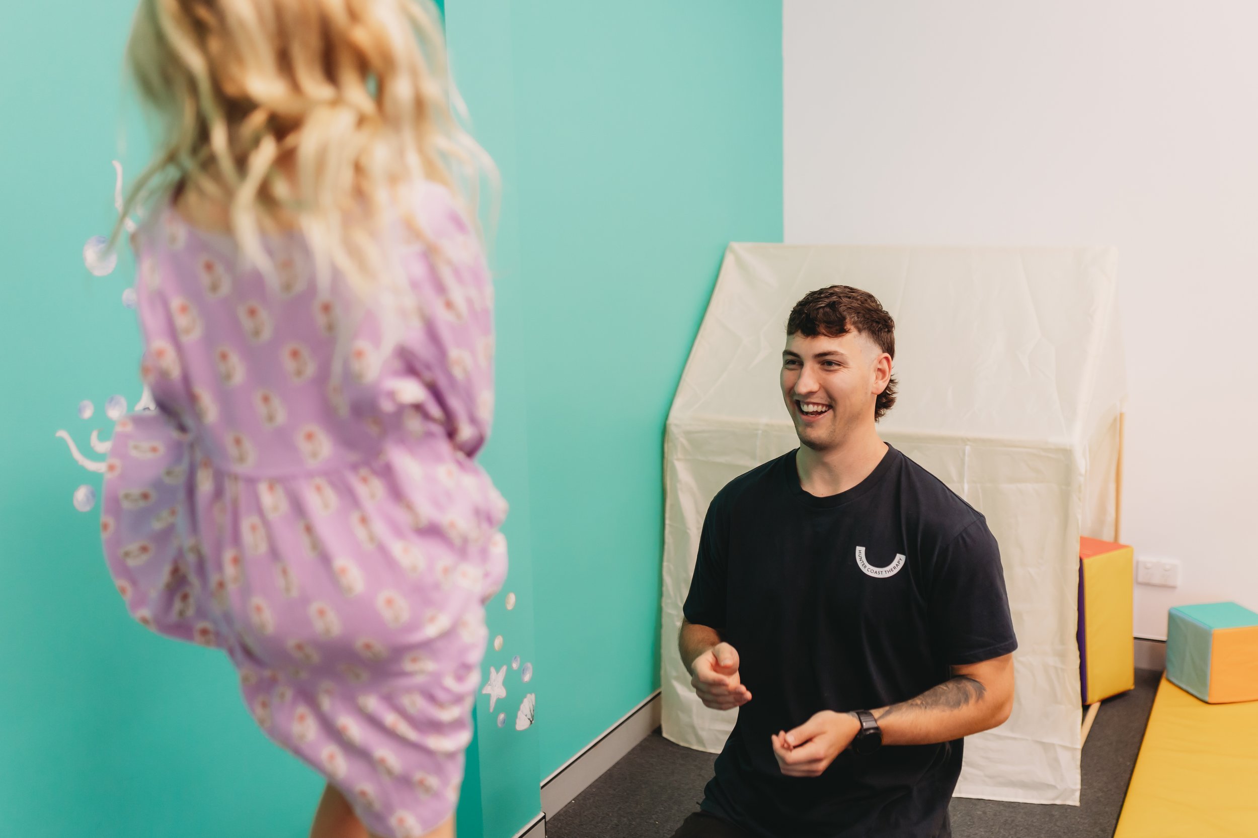 A young man smiling and kneeling in front of a girl on a trampoline in therapy session, in a room with colorful foam blocks and a green wall in Newcasle Australia