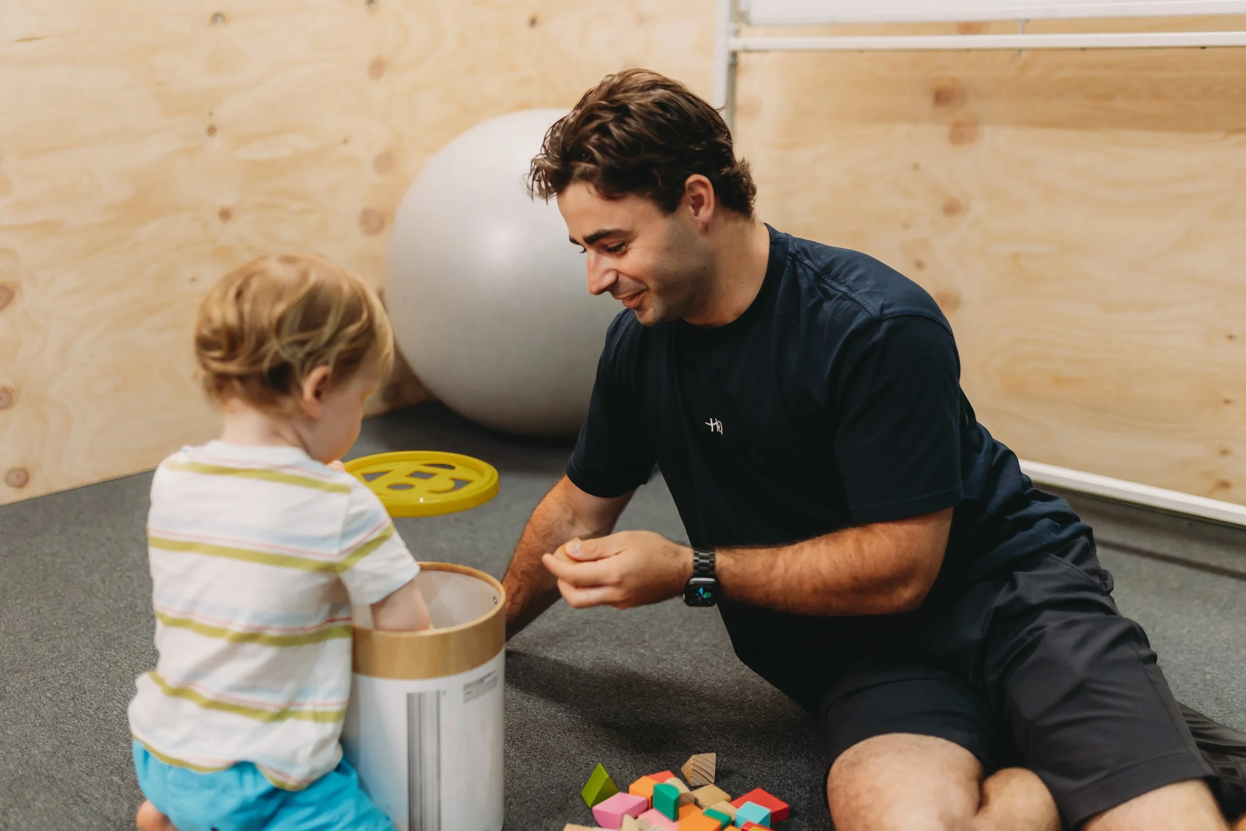 A man and a young child playing with colorful wooden blocks on a carpeted floor in a room with wooden walls and exercise equipment in the background. Therapy session in Newcastle