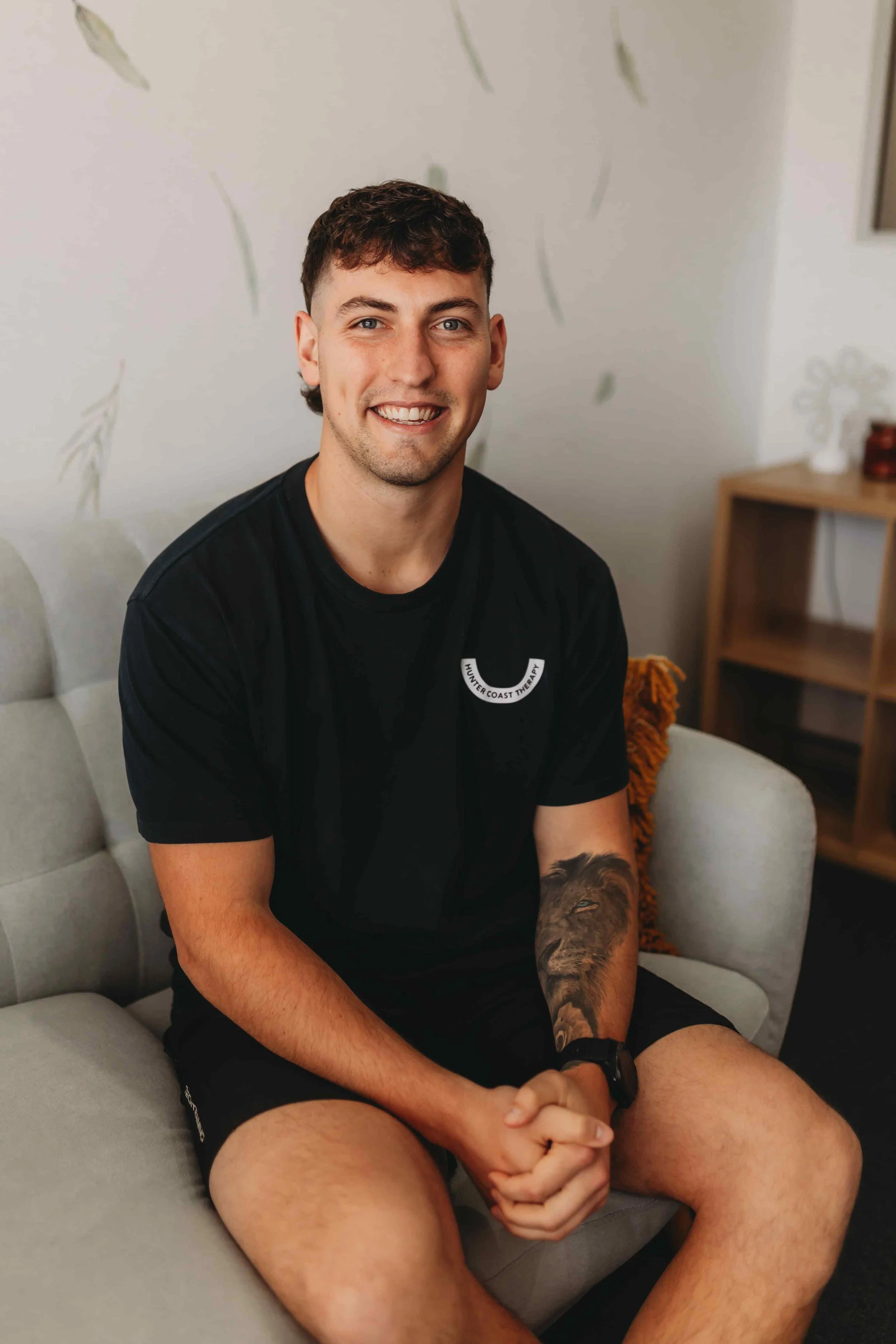 A young man with short curly hair, wearing a black T-shirt with a logo, sitting on a light-colored sofa with his hands clasped, smiling at the camera.