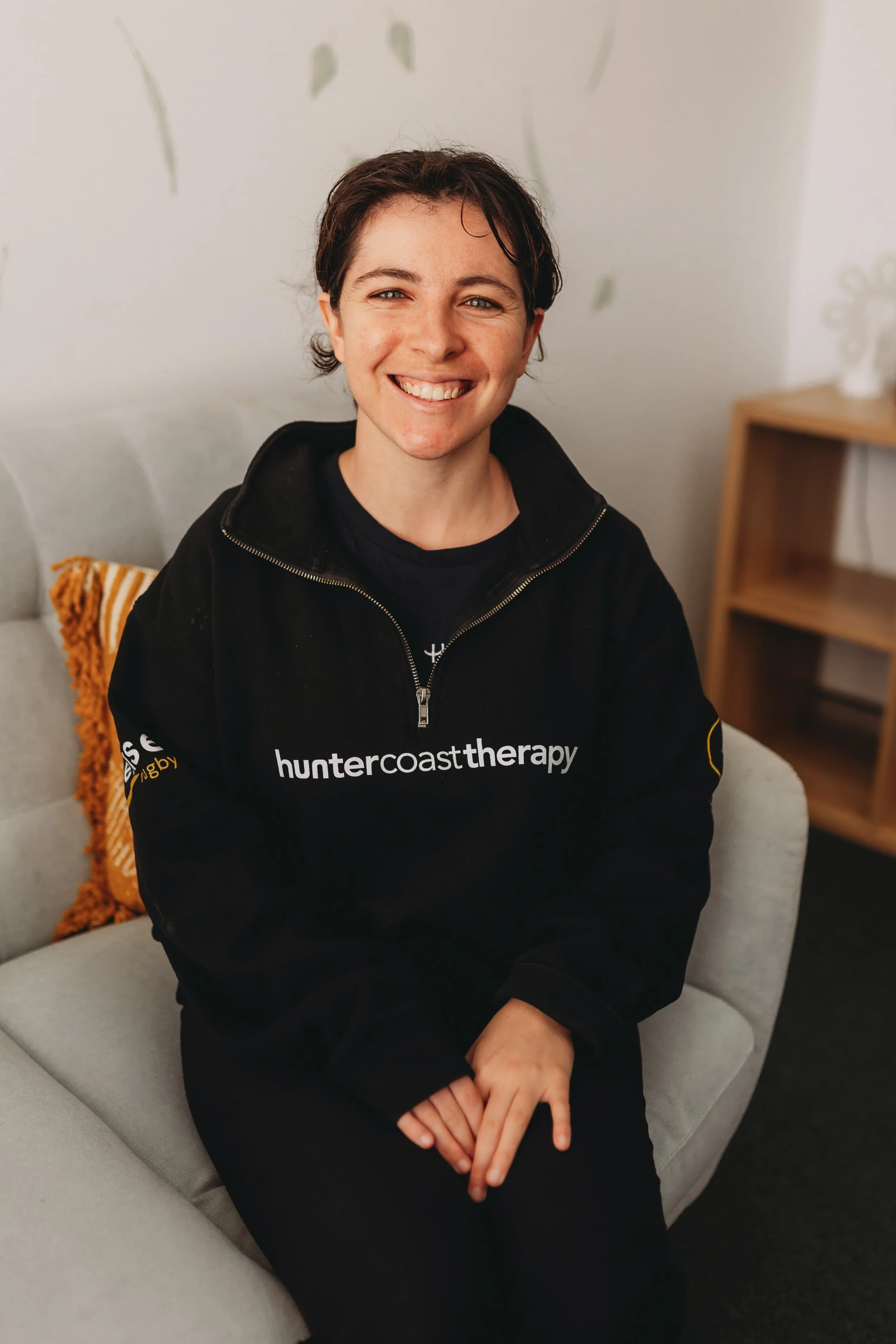 A smiling woman with short dark hair sitting on a light-colored sofa in a room, wearing a black hoodie with 'huntercoasttherapy' written on it.