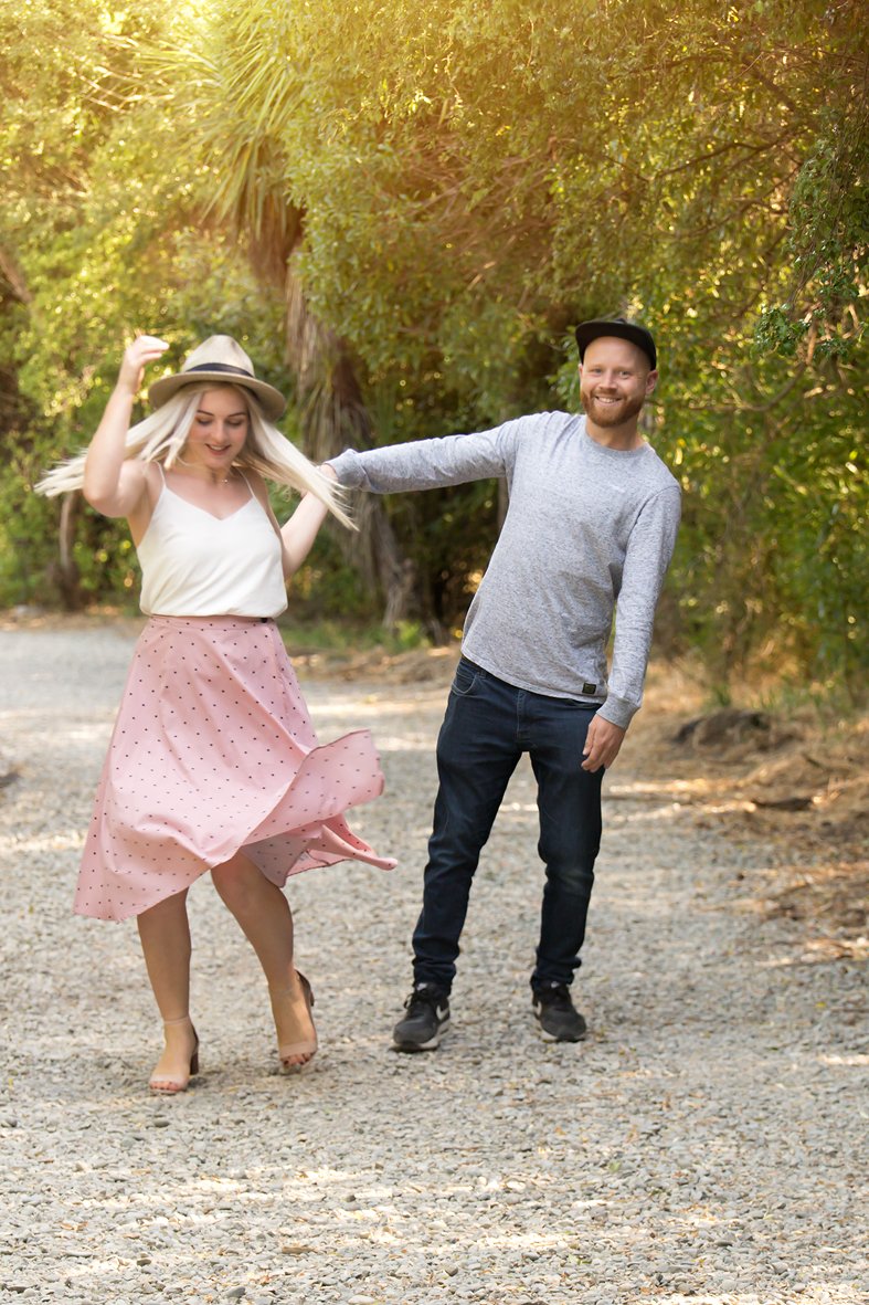 A man and woman are enjoying a playful moment on a dirt path surrounded by trees, the woman is twirling while the man is holding her hand, both smiling.