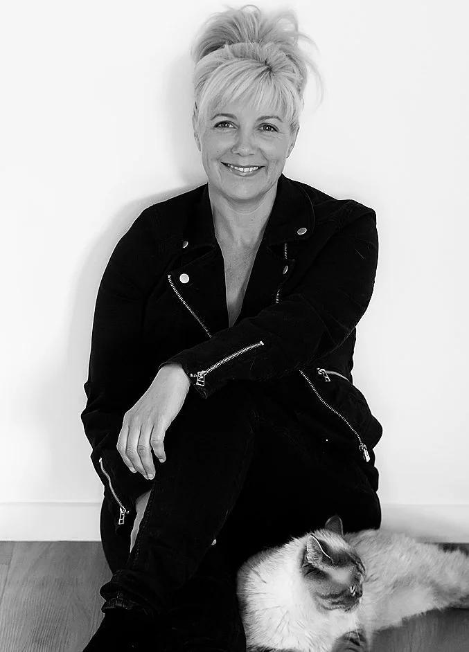 A woman with short, light-colored hair styled in a messy updo, smiling and sitting on the floor. She is wearing a black jacket with zippers. A fluffy cat with long fur is lying beside her on the floor.