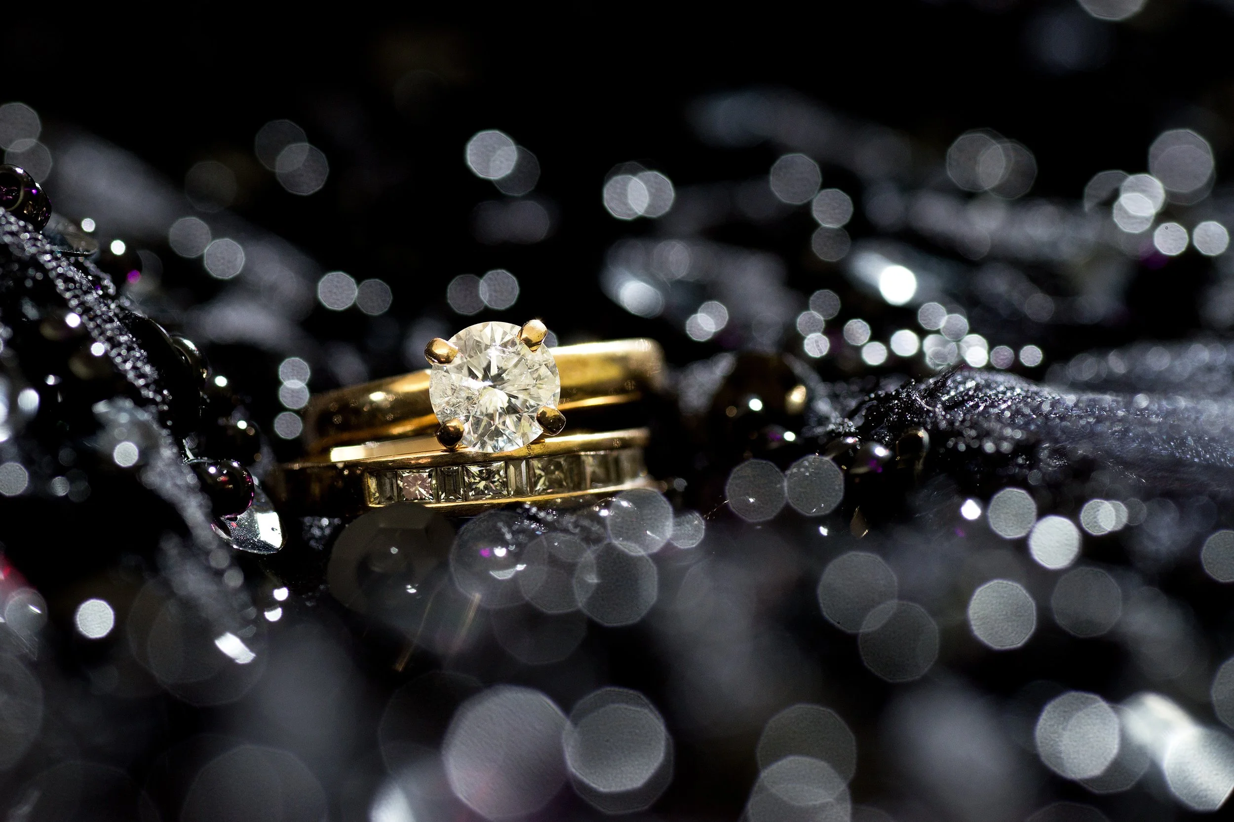 Close-up of a gold ring with a large diamond, resting on a sparkling black surface with scattered jewelry.