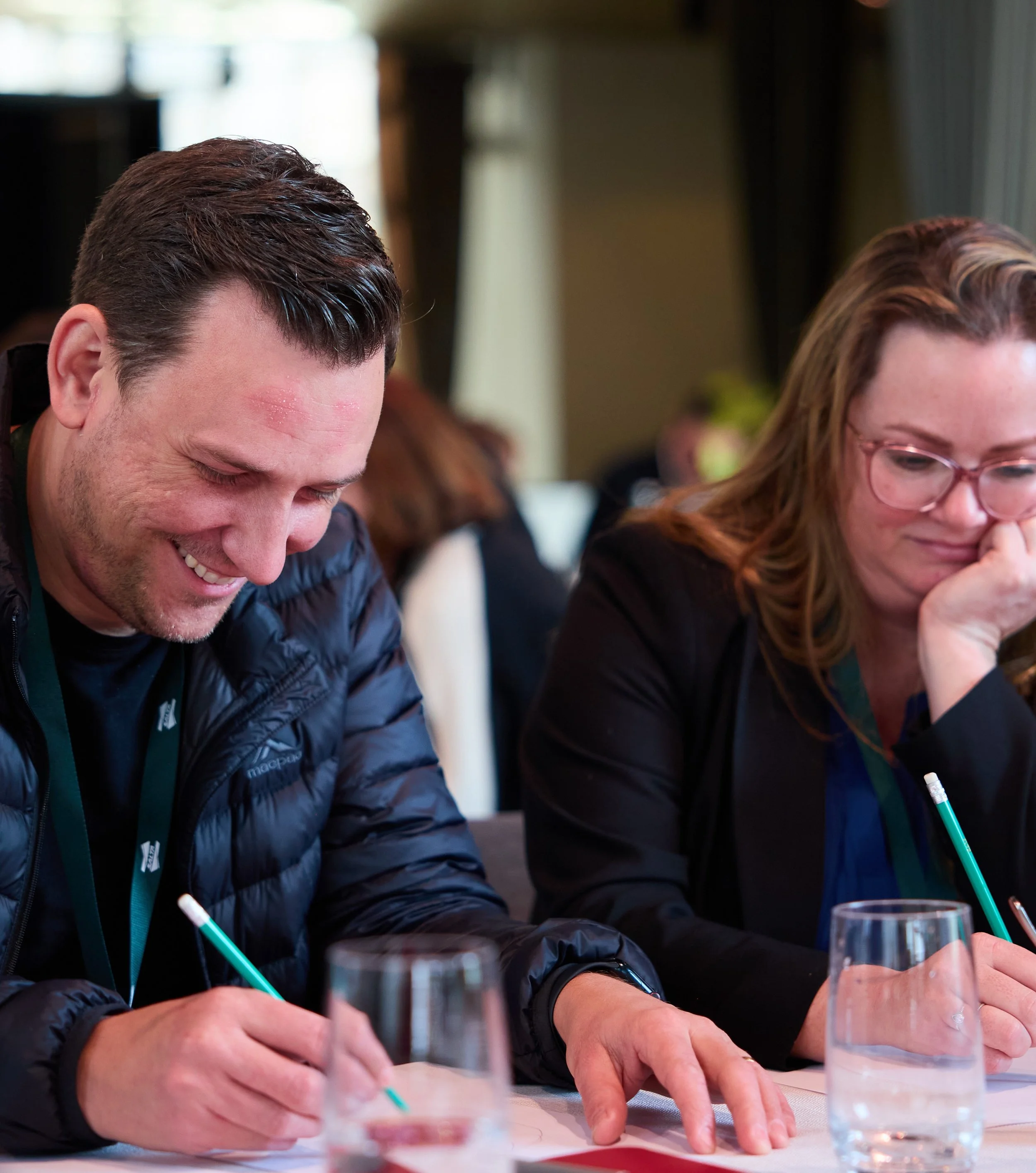 A man with brown hair is writing on paper with a green pen, while a woman with red hair and glasses is sitting next to him, both engaged in their work at a table with glasses of water.