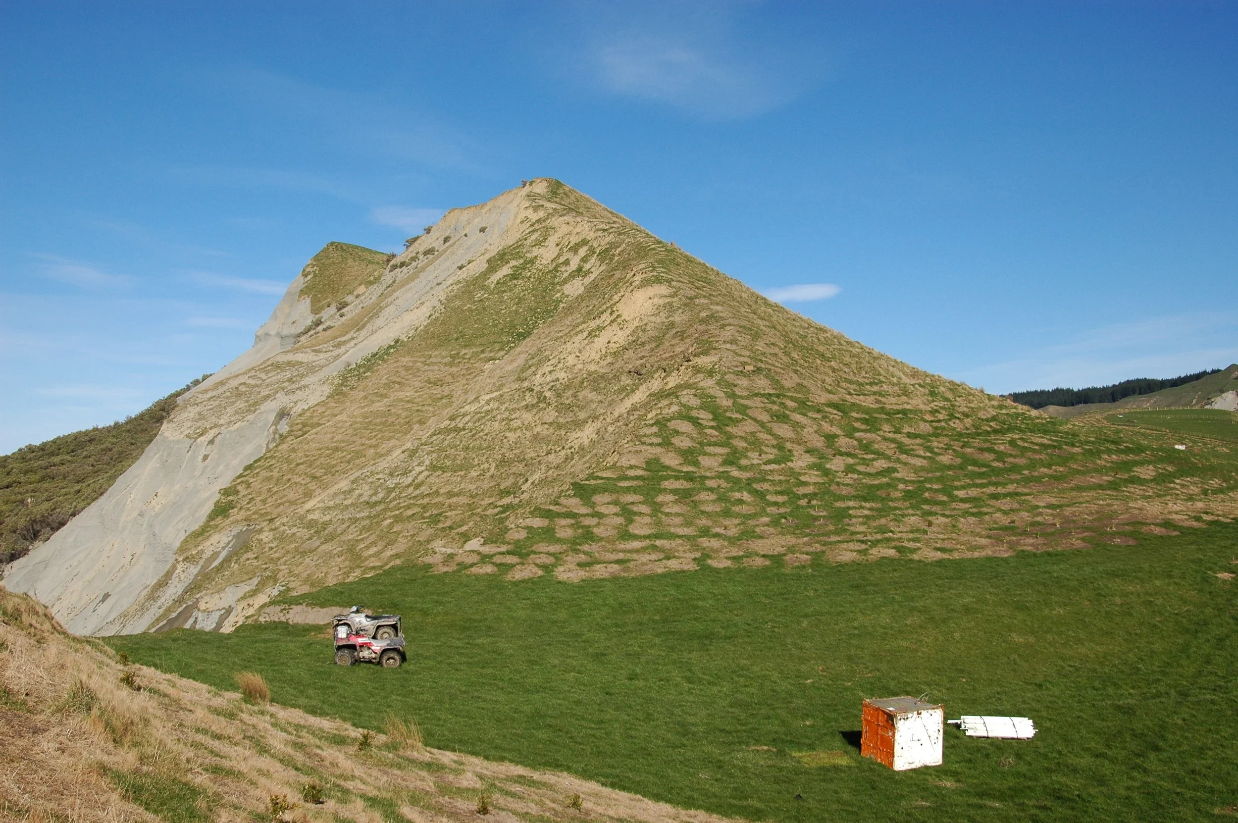 Mountain with grassy slopes and patches of greenery, parked four-wheelers at the base, clear blue sky overhead.