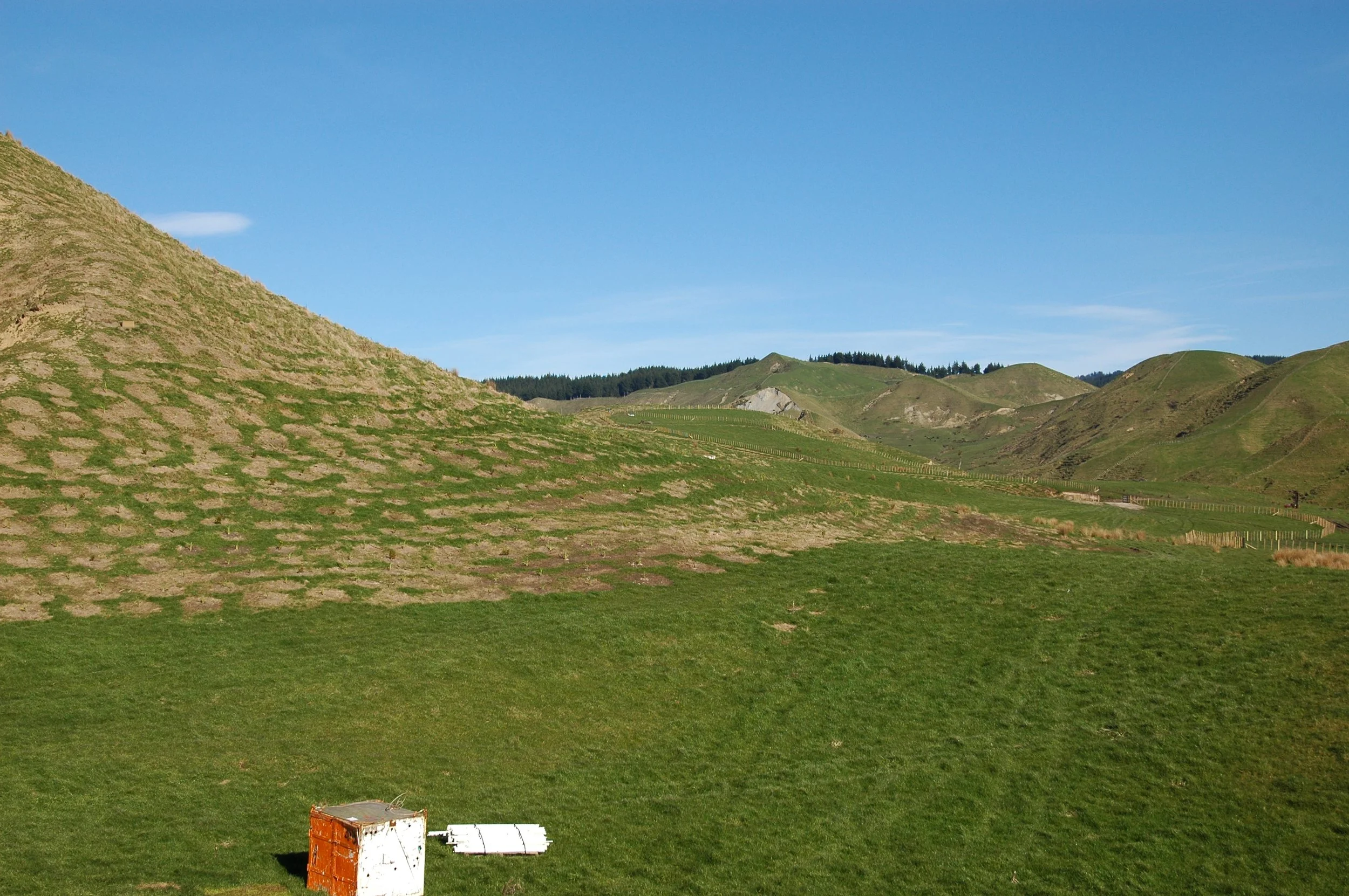 Open grassy field with rolling green hills in the background under a clear blue sky. There is a rusty, white and orange object in the foreground.