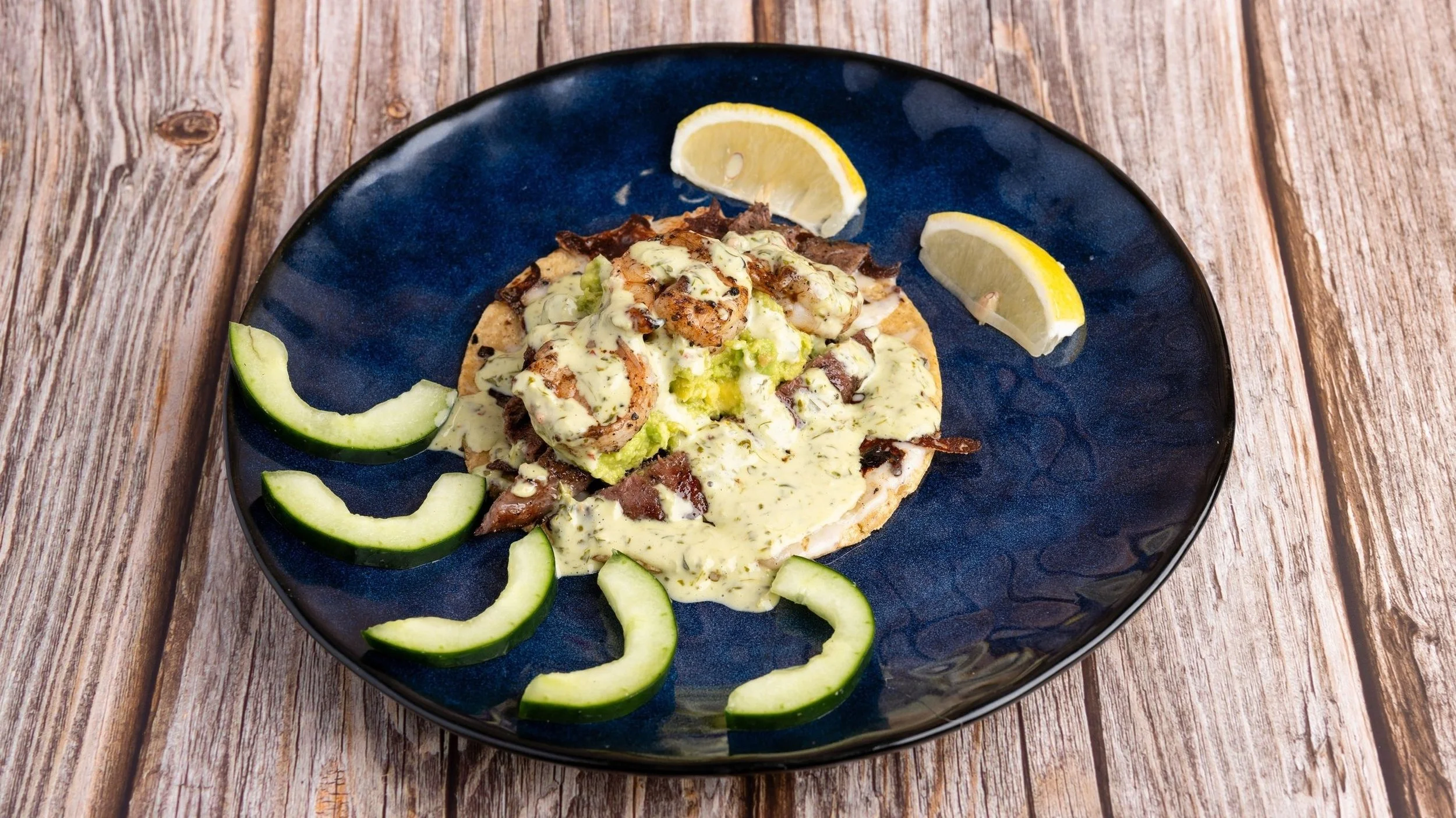 Tacos topped with creamy sauce, garnished with lemon wedges and cucumber slices on a dark blue plate, set on a wooden surface.