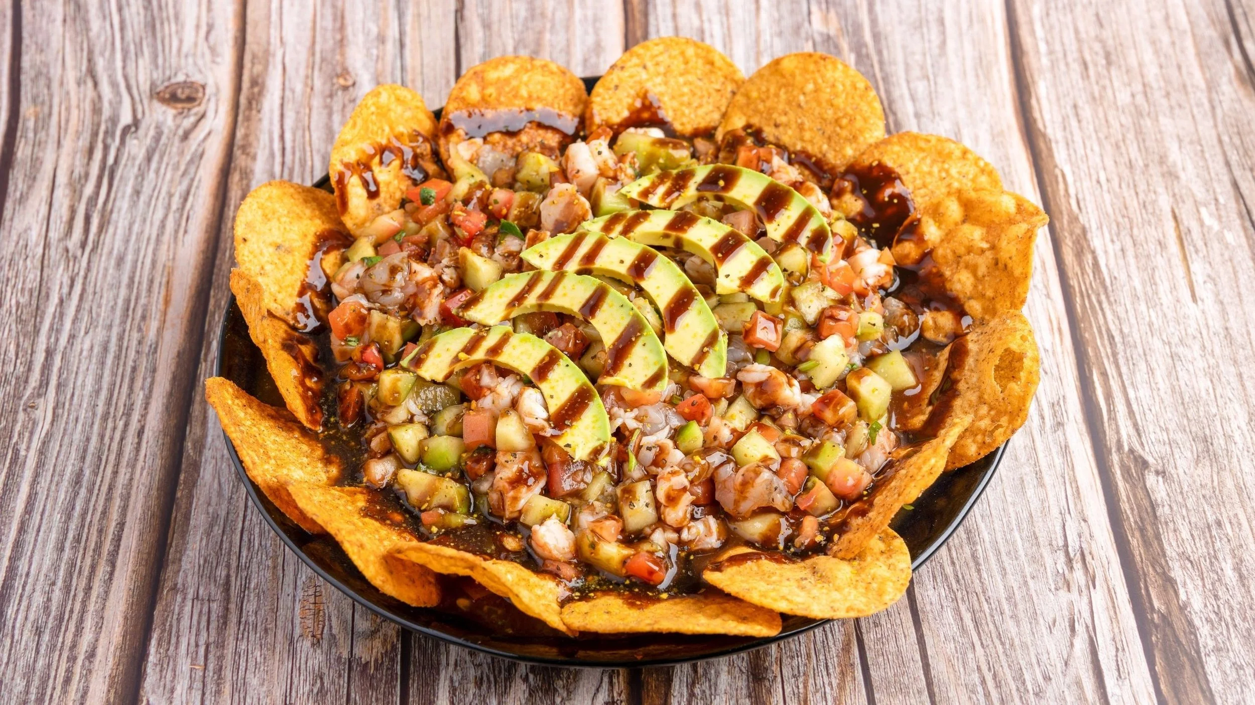 A black bowl filled with nacho chips, topped with a mixture of chopped tomatoes, avocados, and seafood, all drizzled with a dark sauce, placed on a wooden surface.