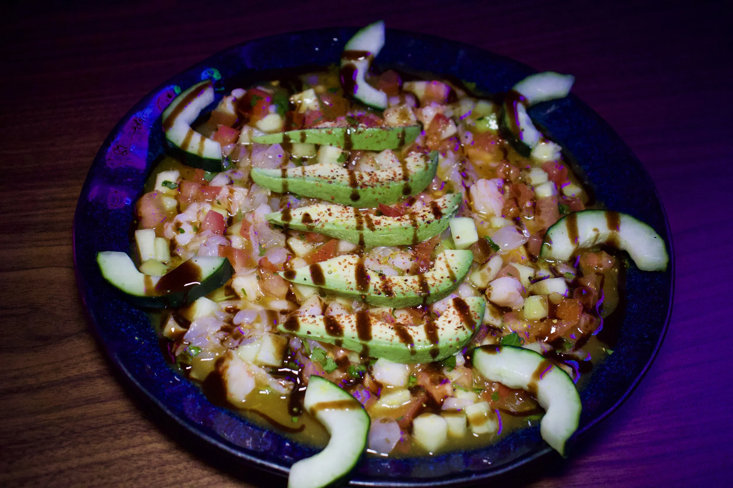 A bowl of chopped vegetables topped with avocado slices, drizzled with dark sauce, on a dark wooden surface.